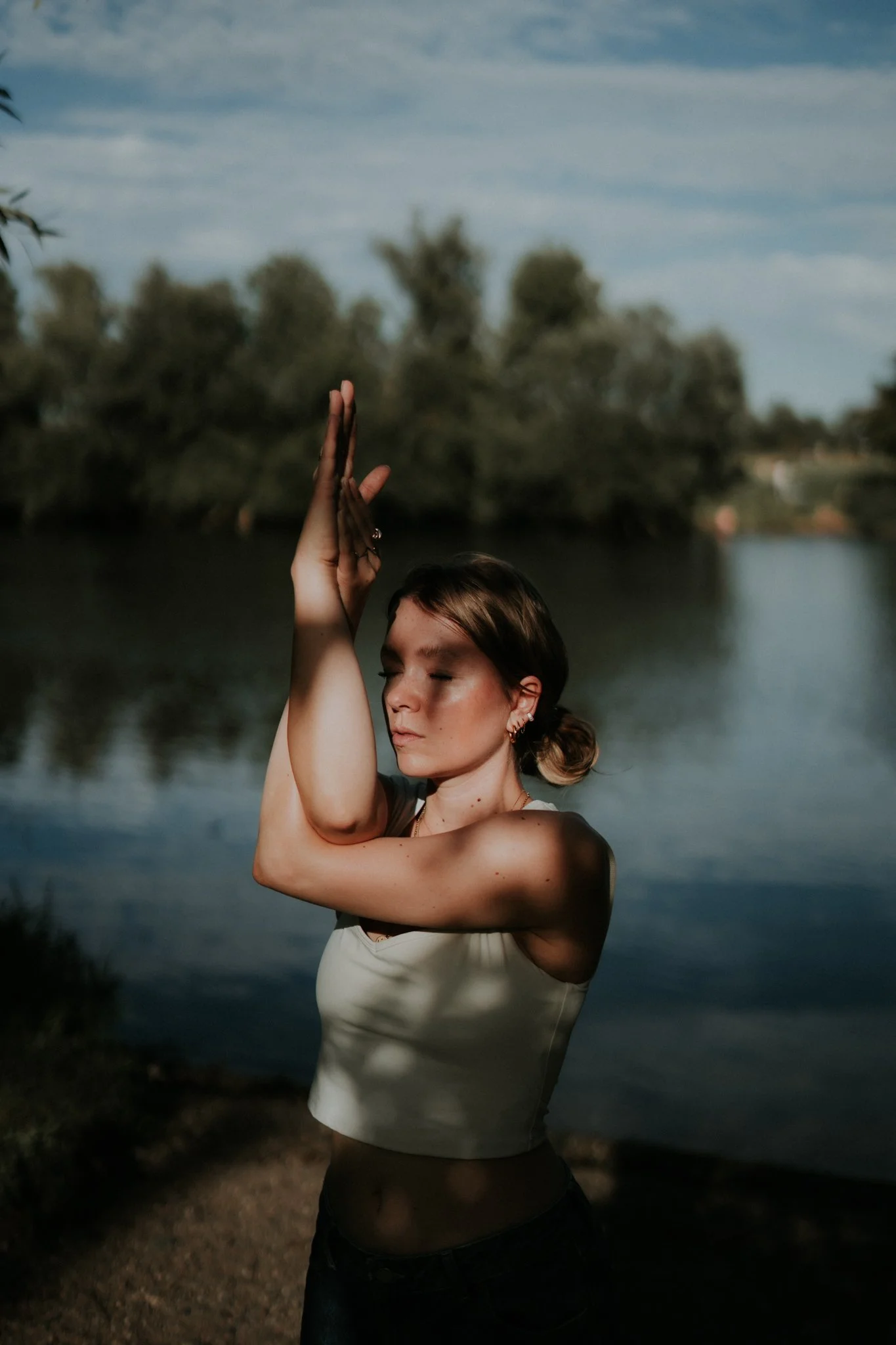 Eine Frau steht mit verschränkten Armen vor einer ruhigen Flusslandschaft unter einem bewölkten Himmel.