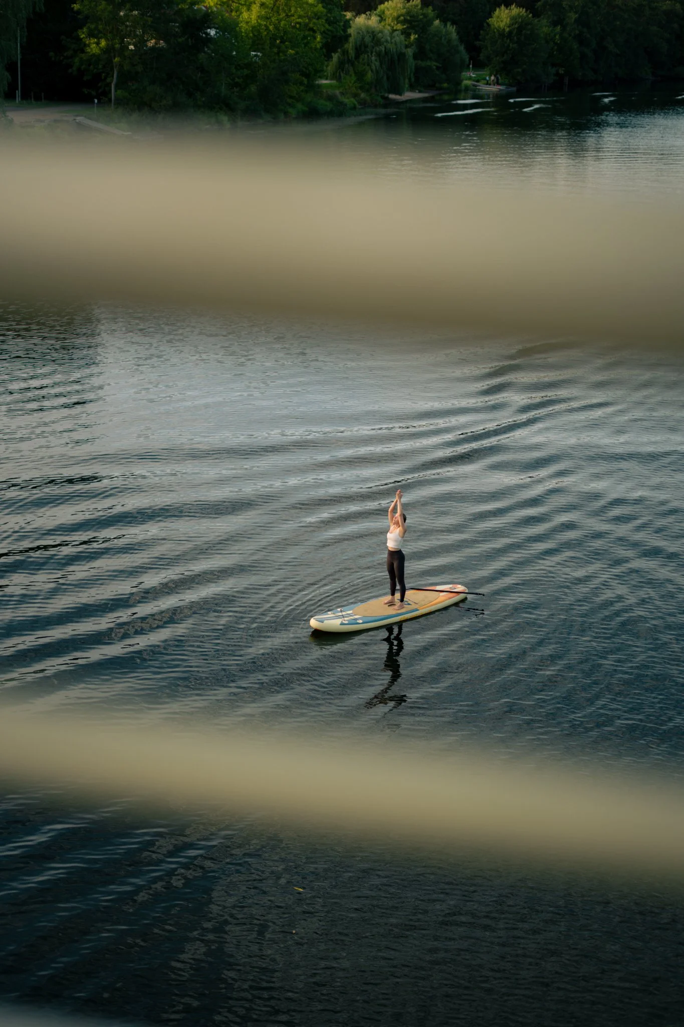 Eine Person steht auf einem Stand-Up-Paddle-Board auf einem ruhigen See in der Dämmerung, umgeben von grünen Bäumen.