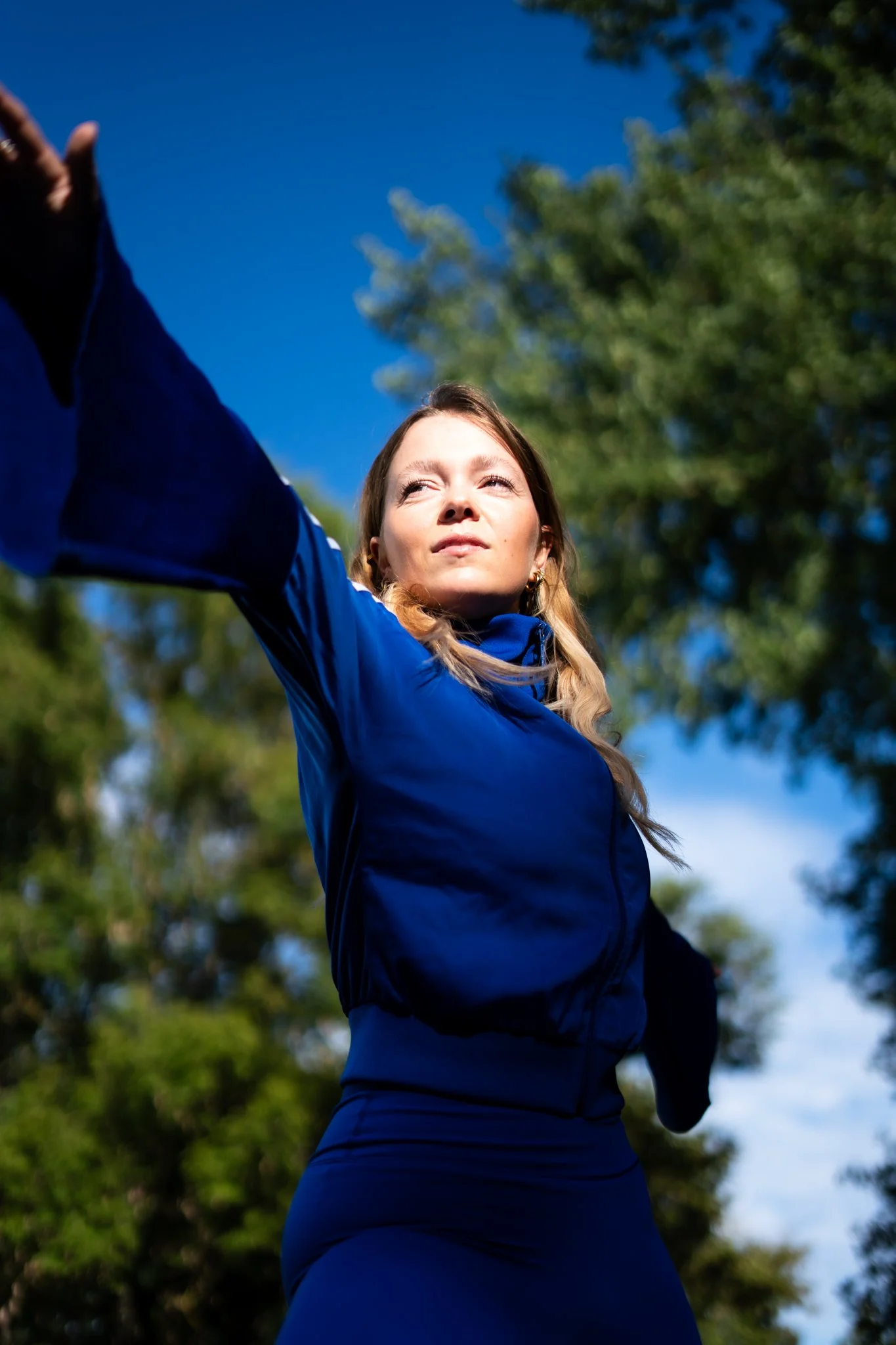Eine Frau in blauer Sportkleidung macht im Freien eine Yoga- oder Meditation-Übung beim Sonnenaufgang oder Sonnenuntergang, umgeben von Bäumen.
