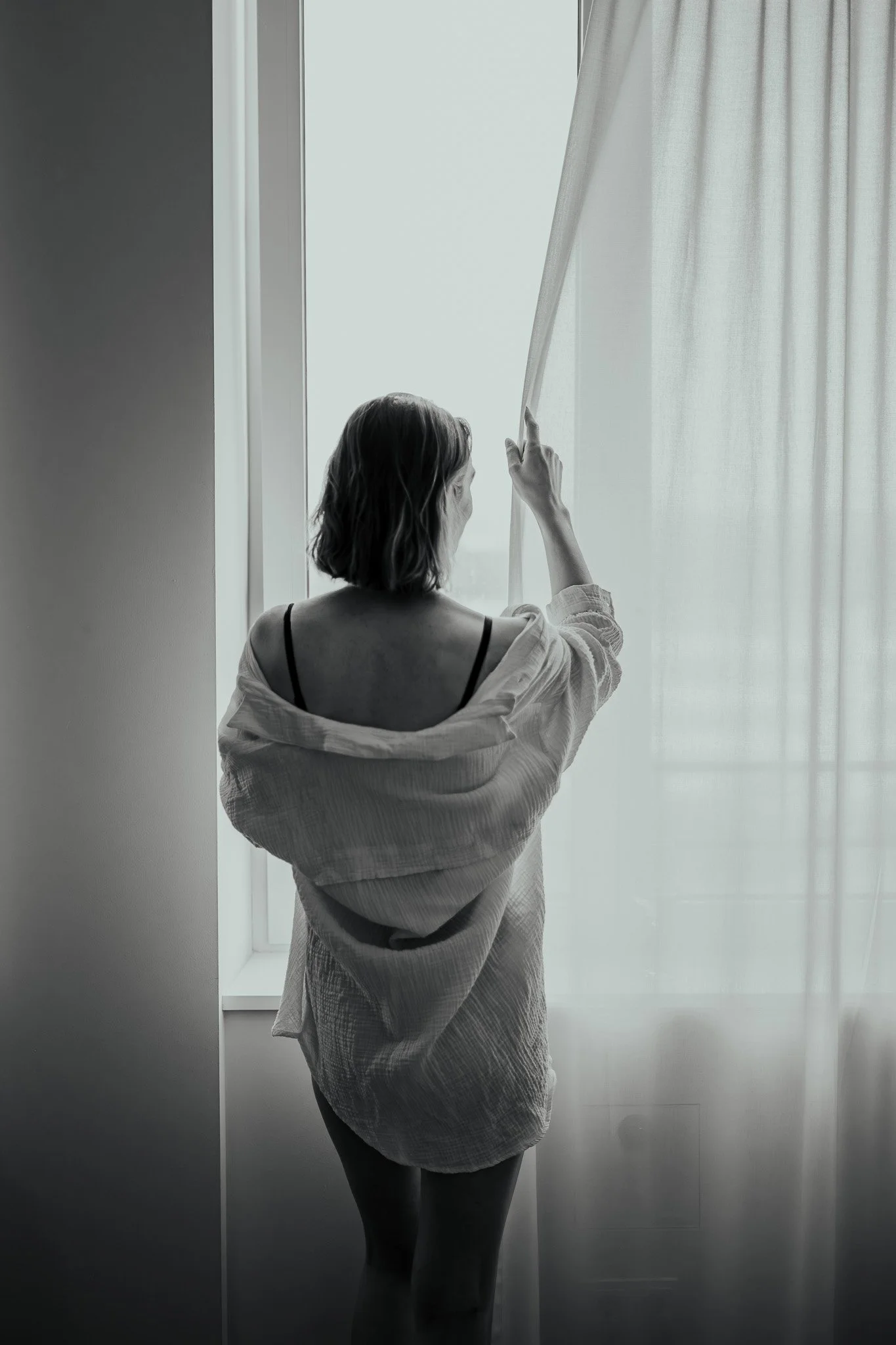 Frau steht vor einem Fenster mit weißen Vorhängen, die sie leicht anhebt, trägt ein off-shoulder Hemd.