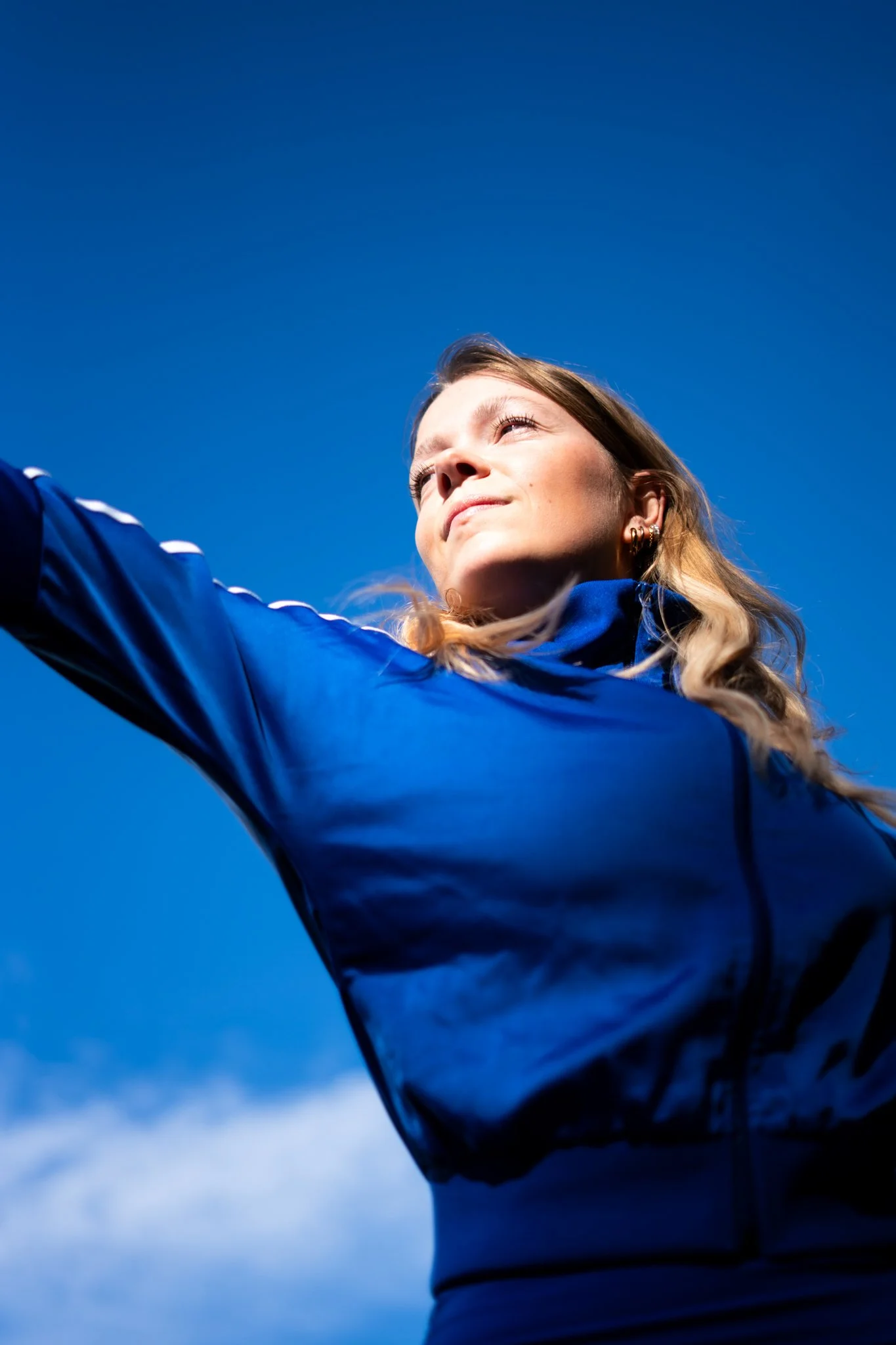 Junge Frau mit langen Haaren, die in der Sonne den Blick nach oben richtet, trägt blaue Sportjacke, blauer Himmel im Hintergrund.