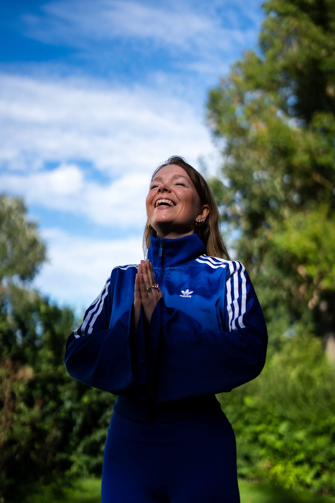 Eine lachende Frau in einer blauen Adidas-Jacke in Namaste-Haltung, draußen in der Natur mit blauen Himmel und grünen Bäumen.