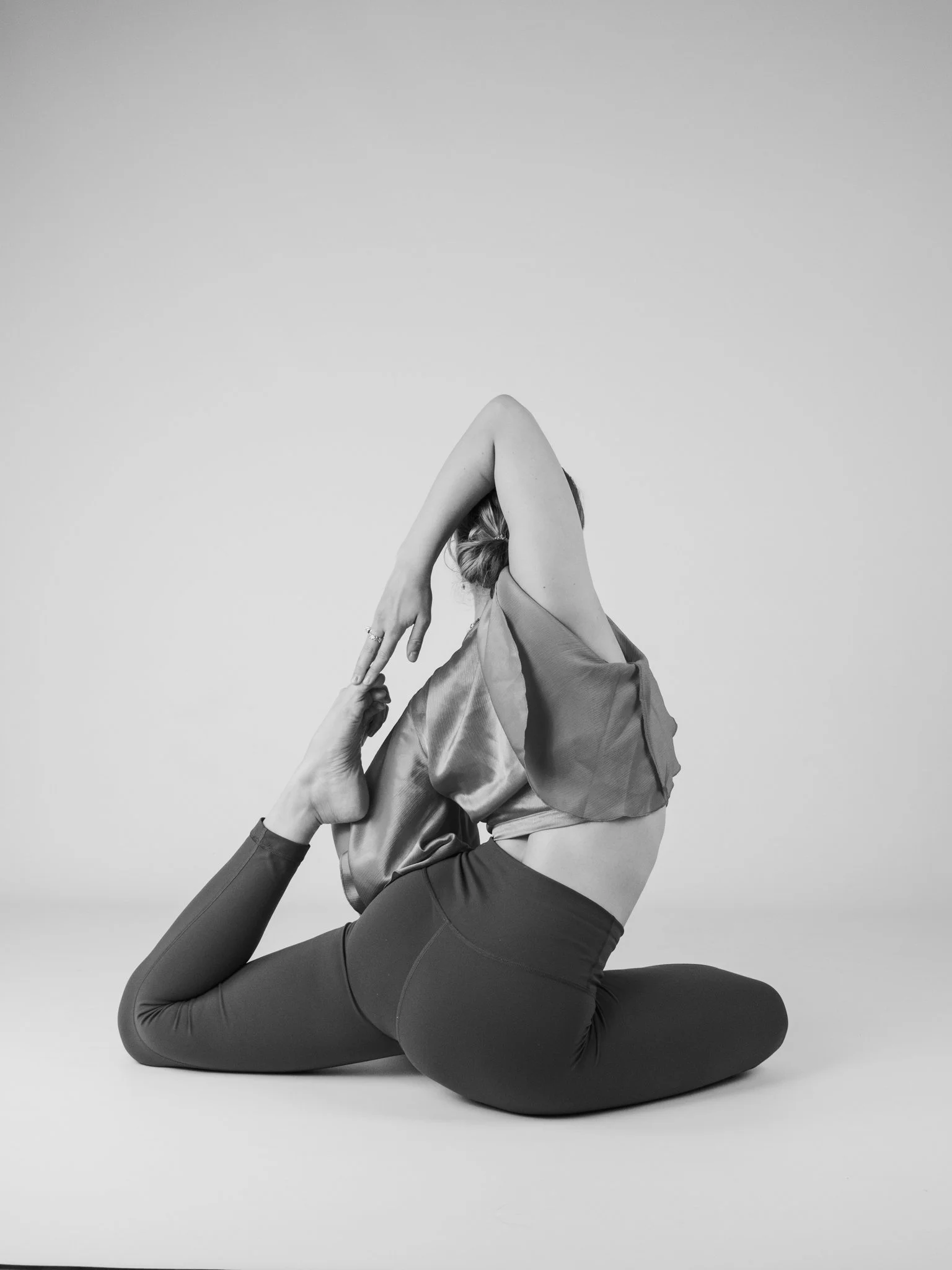Schwarze und weiße, monochrome Fotografie einer Person, die in einer Yoga-Pose sitzt, die Beine gekreuzt und die Arme über den Kopf gestreckt hat, während sie auf dem Boden sitzt.