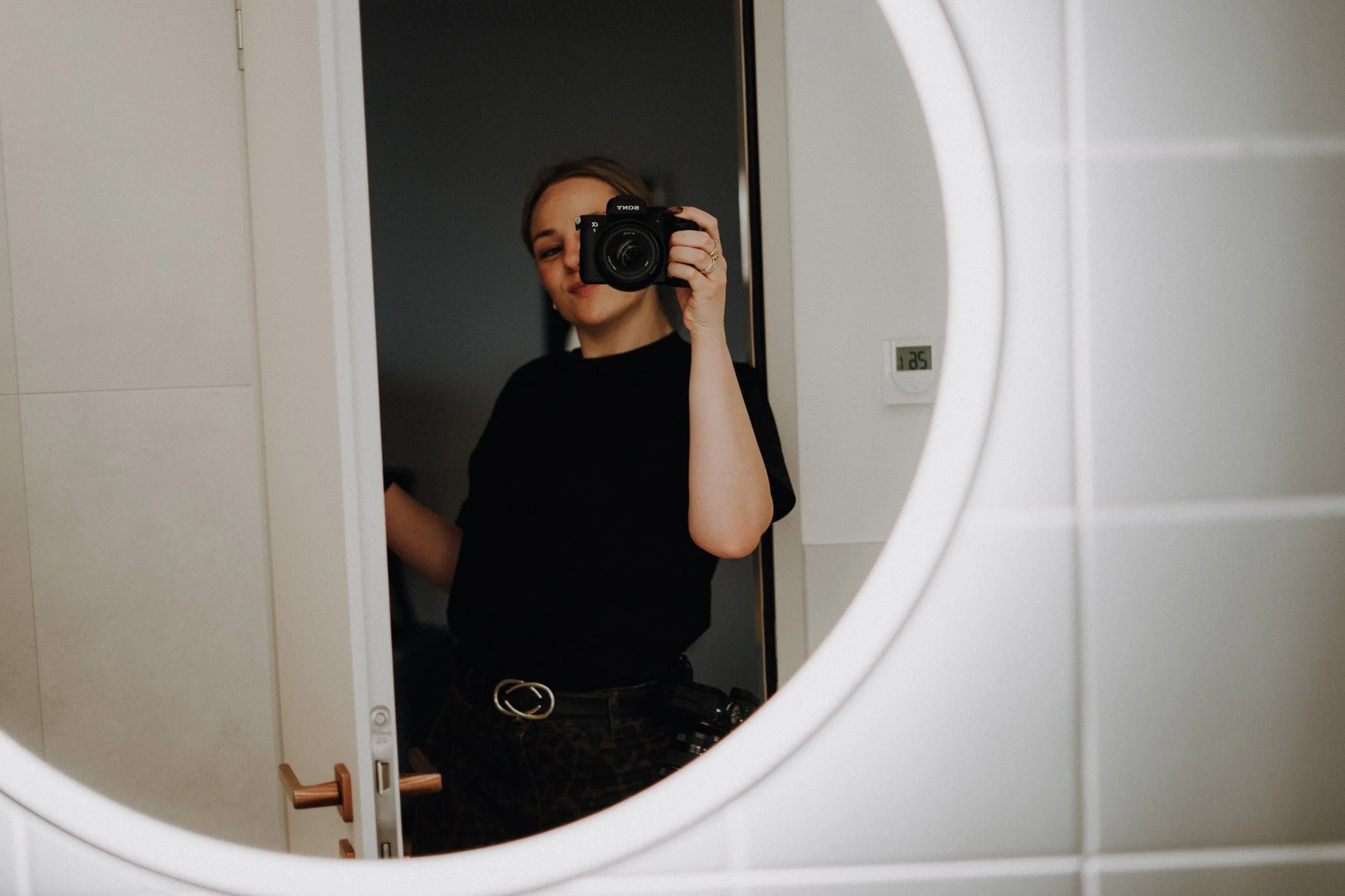 Eine junge Frau macht ein Selfie vor einem Spiegel in einem Badezimmer, die Kamera verdeckt ihr Gesicht, sie trägt einen schwarzen T-Shirt und schwarze Hose mit Gürtel.