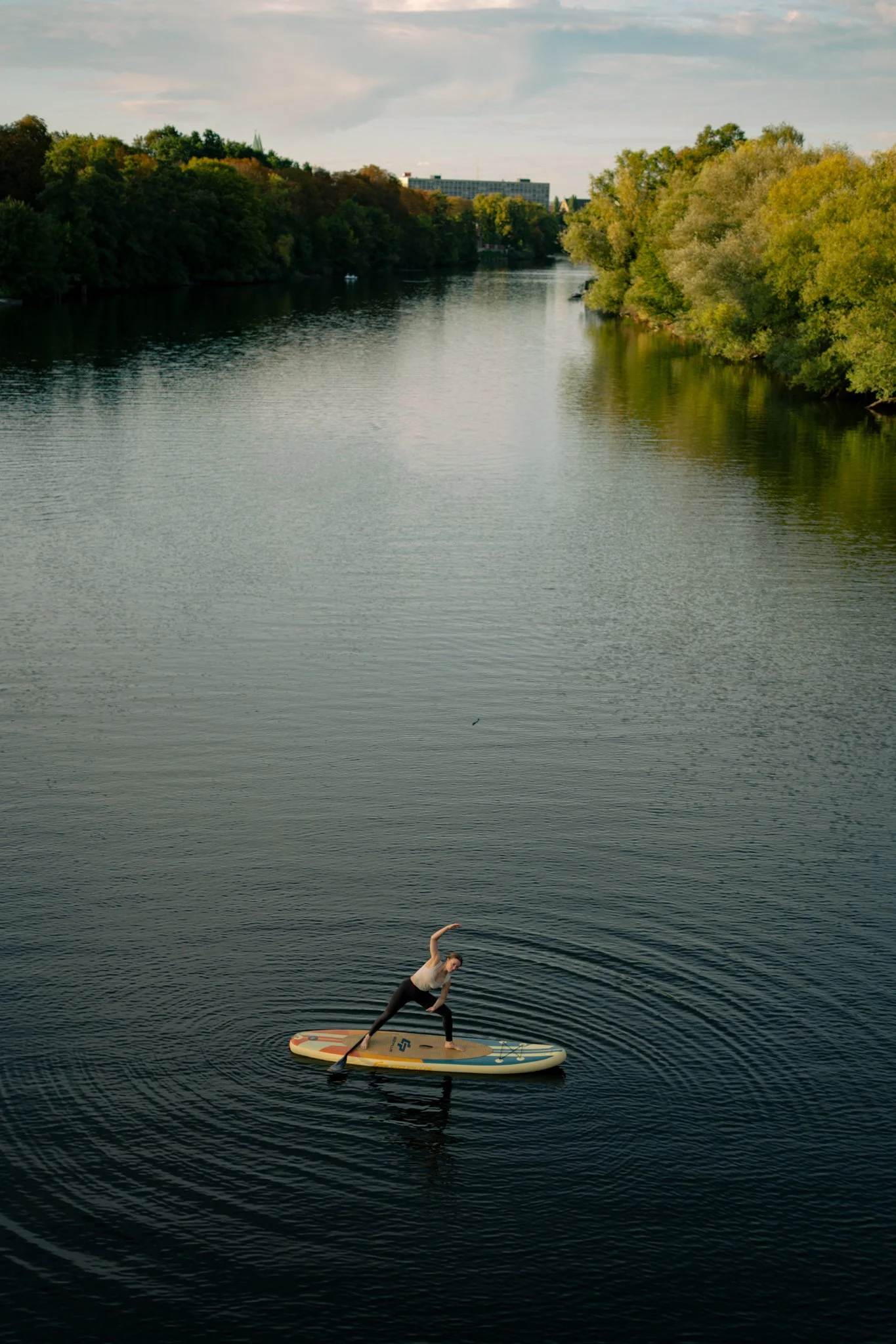 Eine Frau beim Yoga auf einem Stand-Up-Paddle-Board auf einem Fluss, umgeben von Bäumen und Gebäuden im Hintergrund.