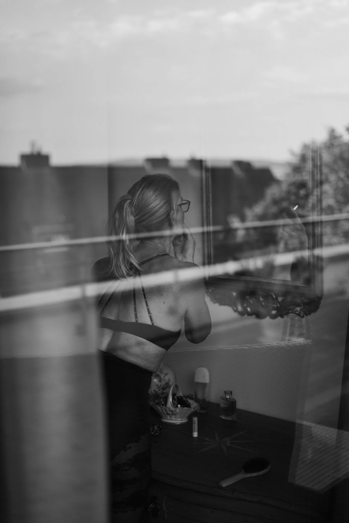 Eine Frau trägt einen schwarz-weißen BH und schminkt sich in einem Raum, der durch eine Glasscheibe gesehen wird.