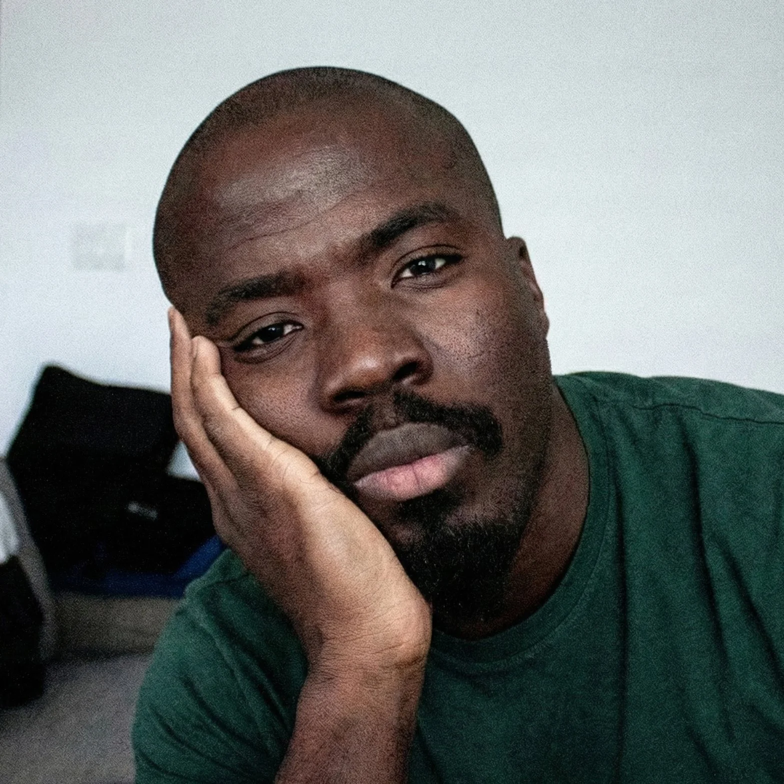 A close-up of a man resting his head on his hand, looking into the camera with a serious expression, wearing a green shirt, with a white wall in the background.