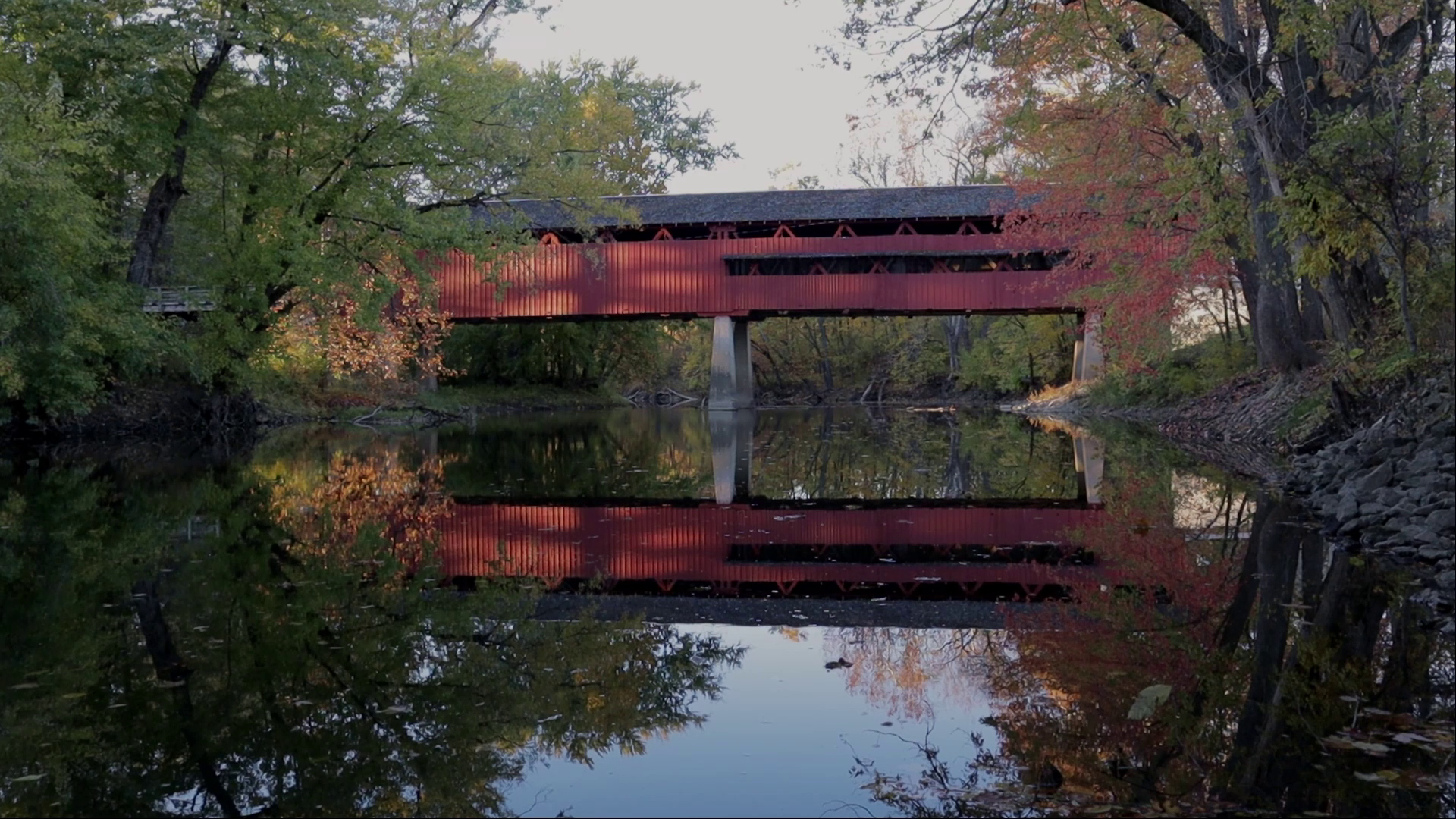 A red covered bridge spans across a calm river, surrounded by trees with autumn foliage, reflecting in the water.