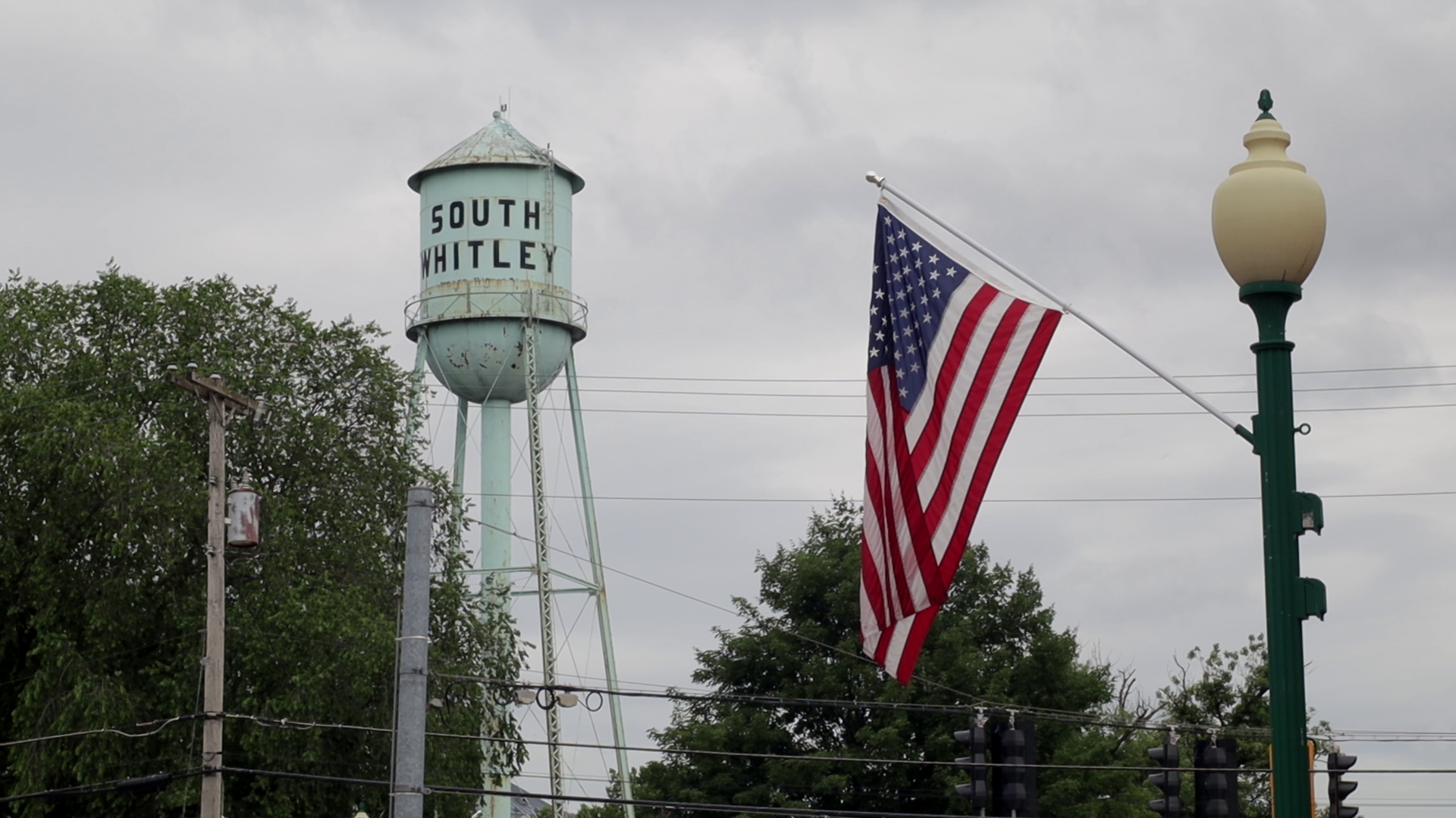 A water tower labeled 'South Whitley', an American flag, a street lamp, and utility poles with power lines under a cloudy sky.