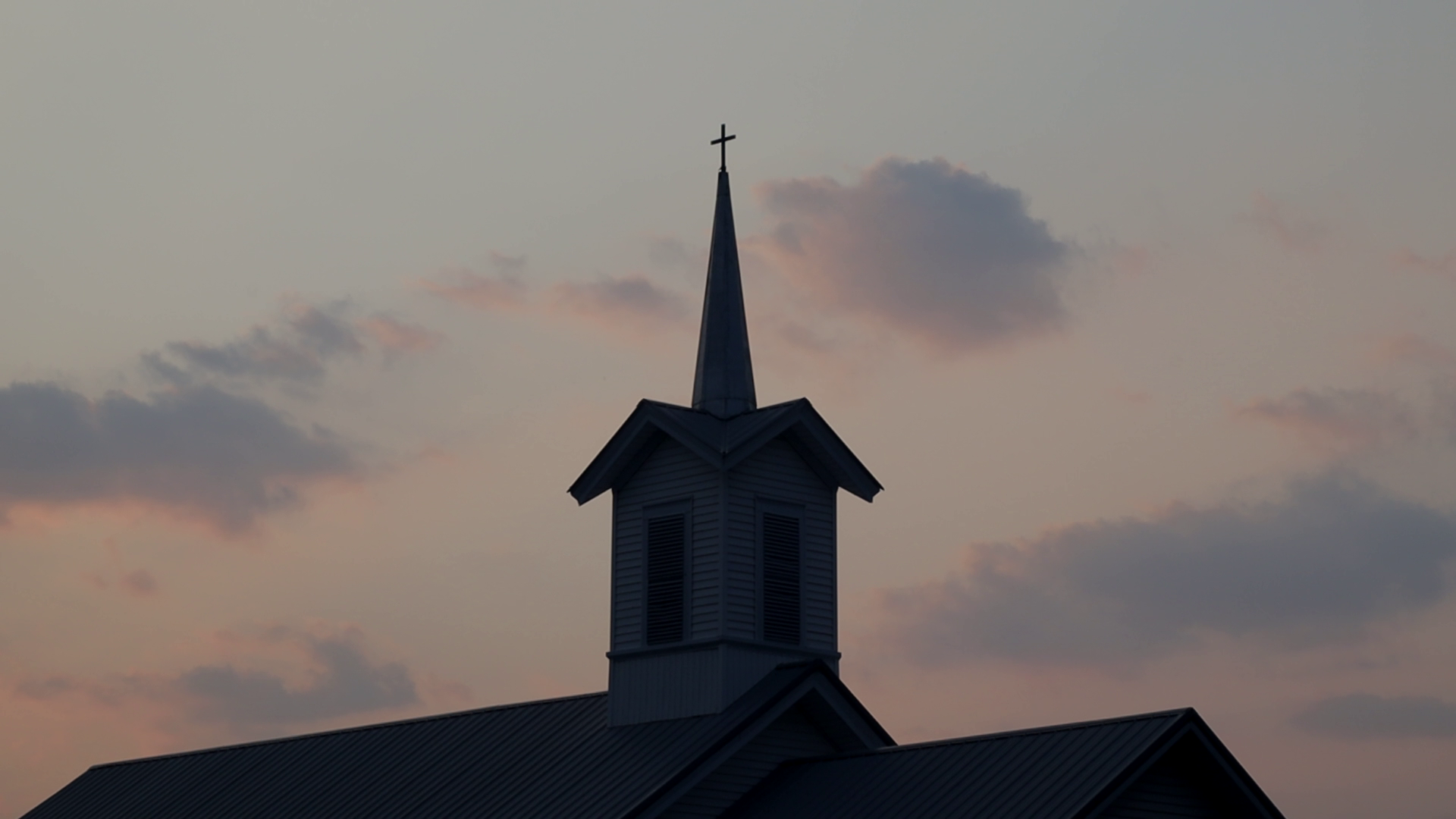 A church steeple with a cross on top against a sunset sky with clouds.