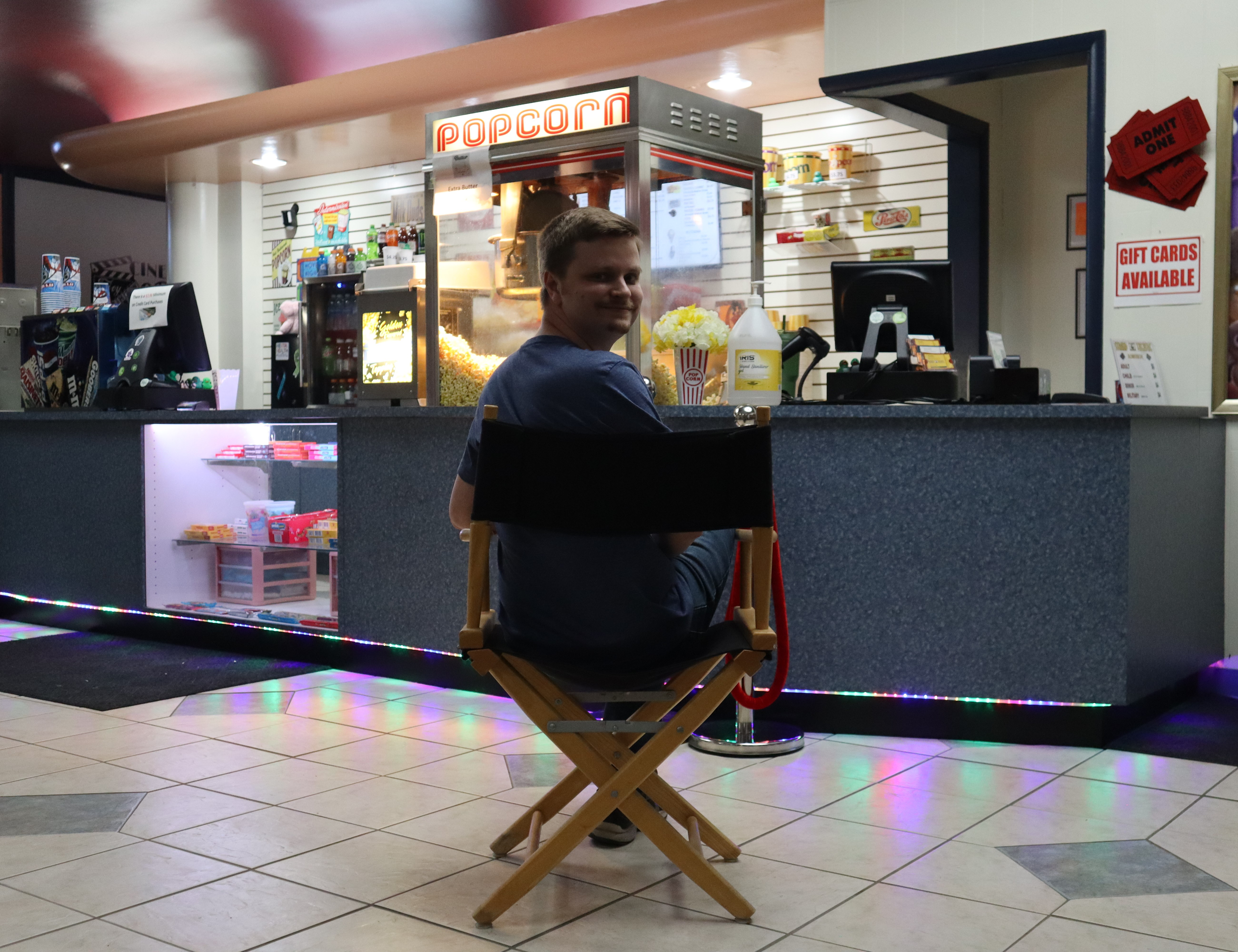 A young man sitting on a black director's chair at a popcorn stand in a shopping mall, with colorful LED lights under the counter, and a small display case with snacks in front.