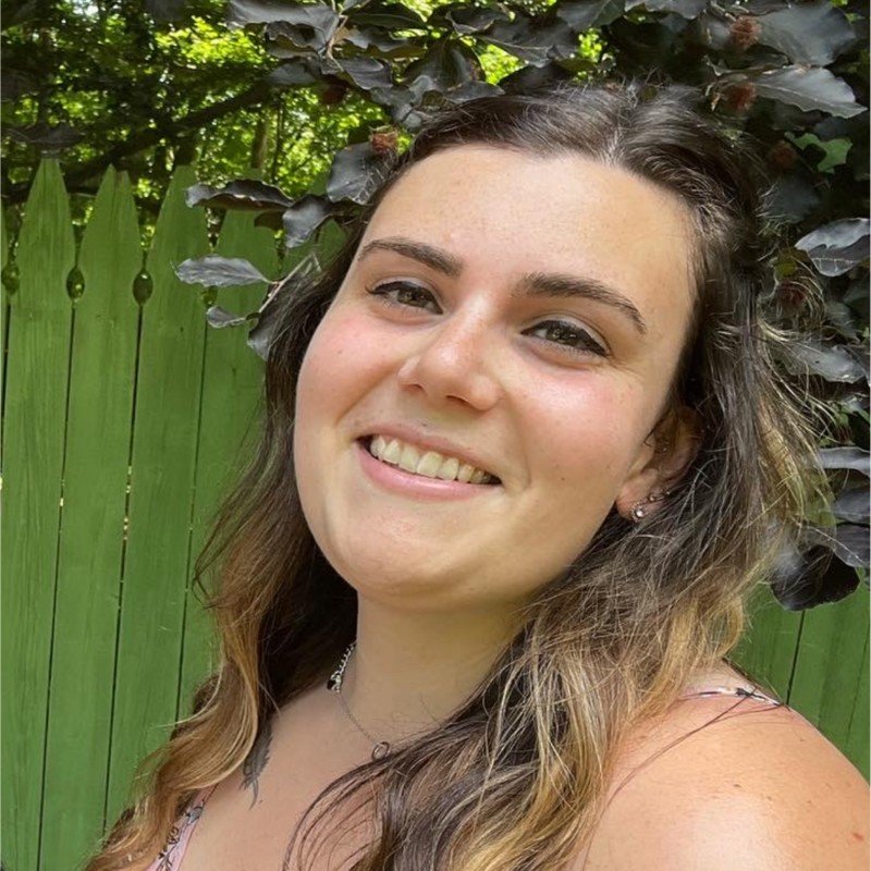 A young woman smiling outdoors with a wooden fence and green foliage in the background.