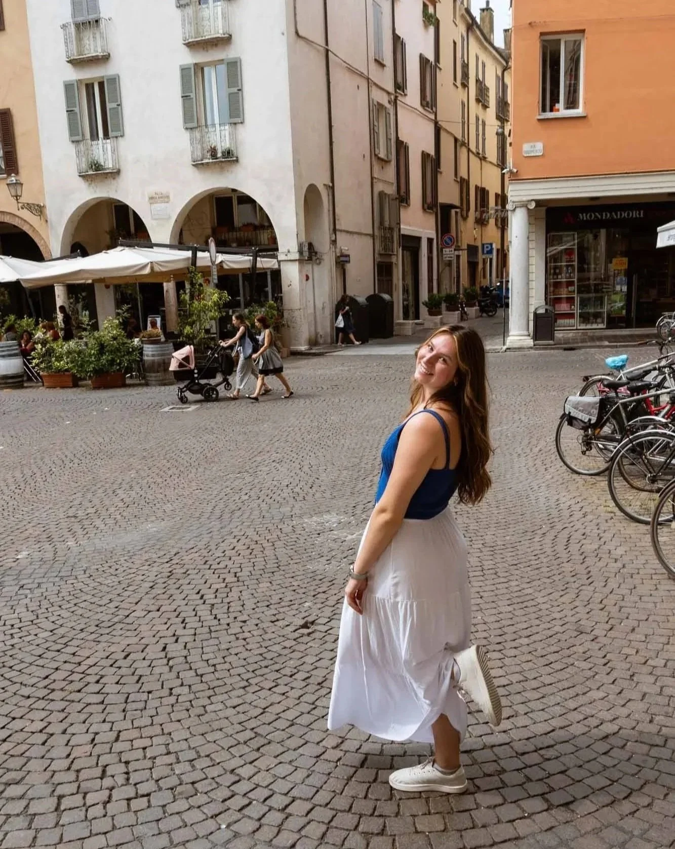 A woman with long brown hair smiling and dressed in a blue tank top and white skirt, standing on cobblestone street in a European town, with bikes parked to the right and a restaurant with outdoor seating to the left.