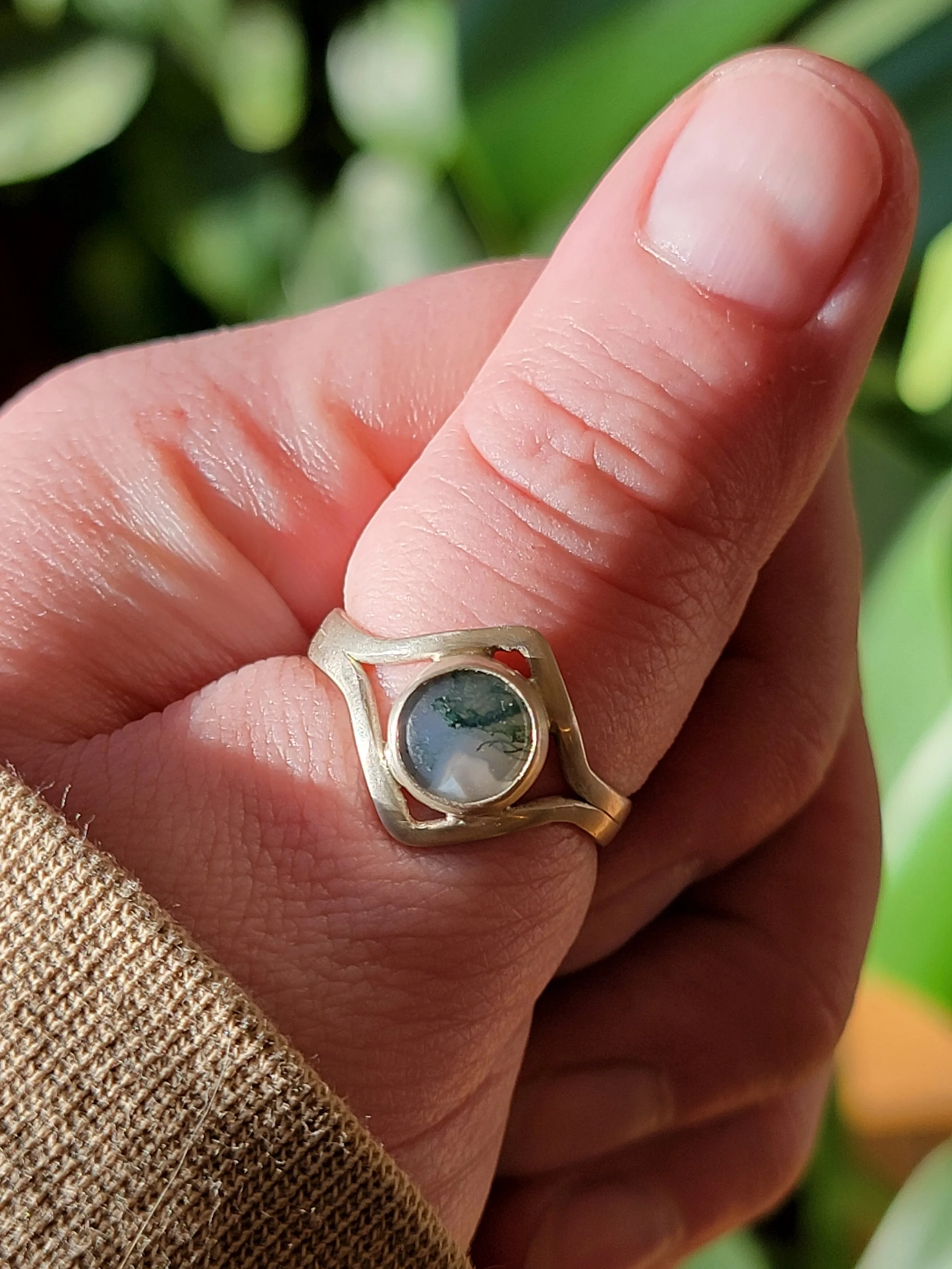 A hand wearing a gold ring with a rounded, semi-transparent gemstone, against a blurred green background.