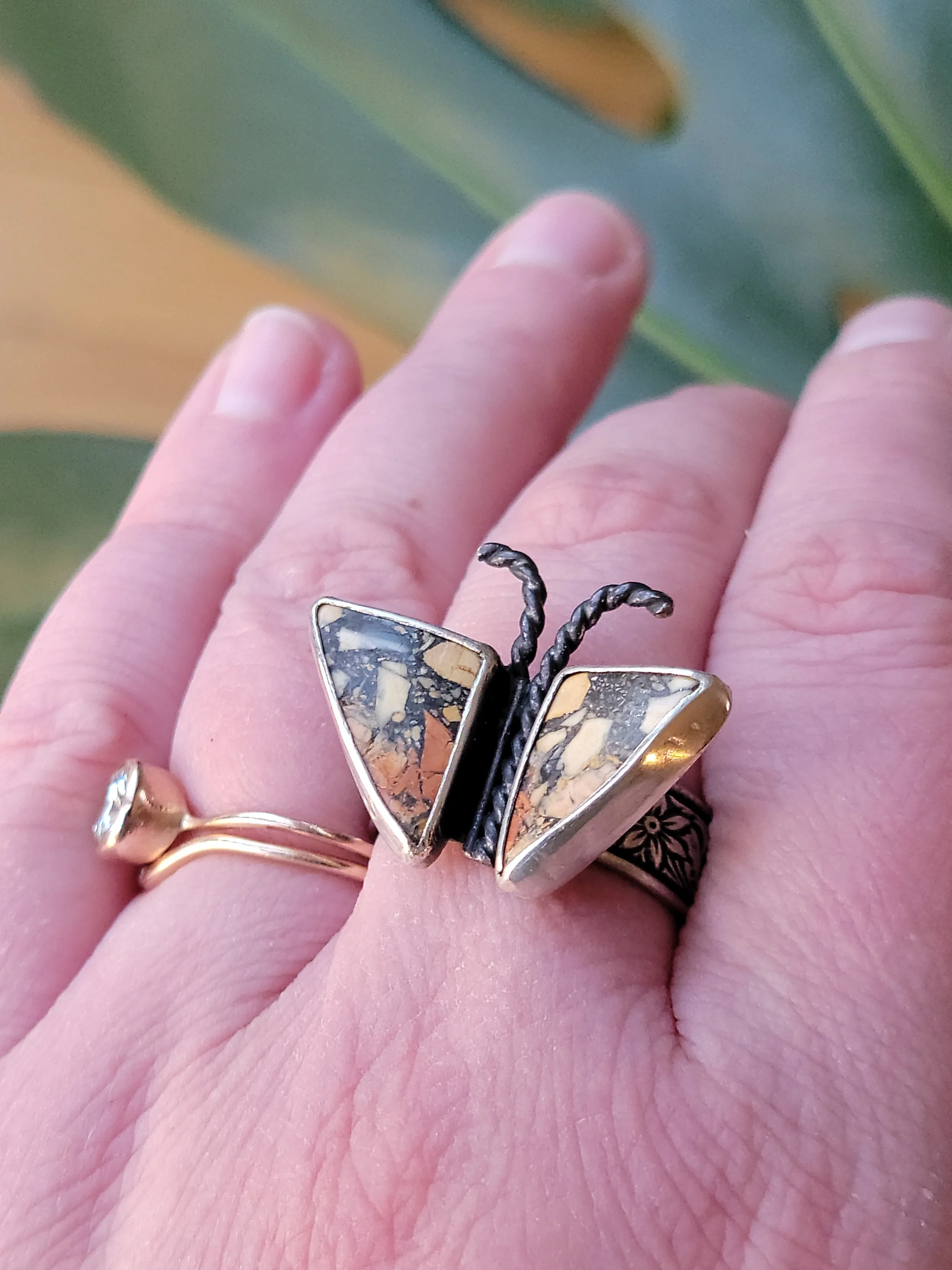 Close-up of a person's hand wearing a butterfly-shaped ring with black and beige marbled wings and twisted black antennae, with a gold band ring featuring a small gemstone, against a background of blurred green leaves.