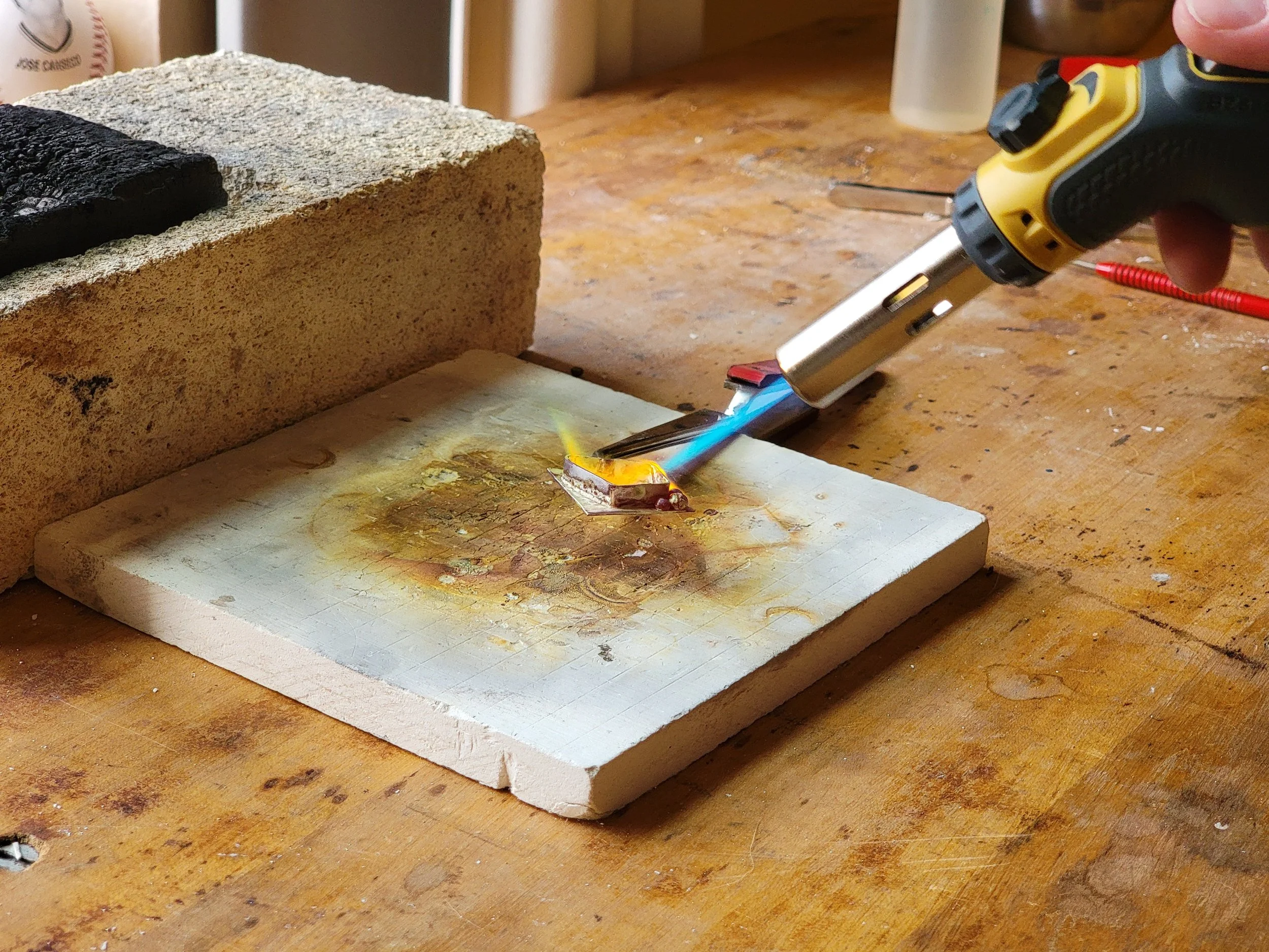 A person uses a soldering iron with a blue flame to work on a small electronic component placed on a ceramic tile, with a block of mineral insulating material in the background on a wooden workbench.