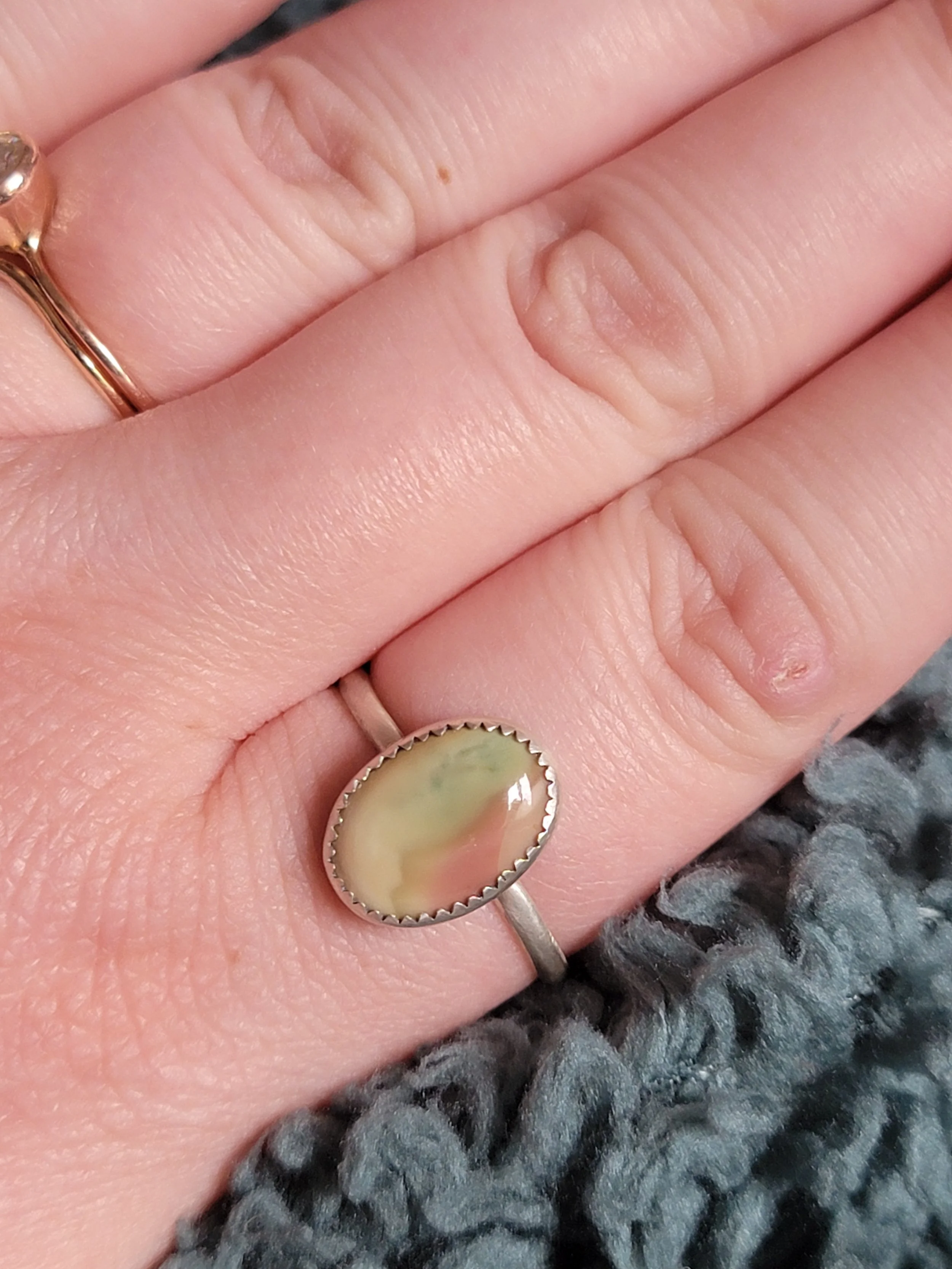 Close-up of a hand with two rings, one on the middle finger and one on the ring finger. The ring on the ring finger is an oval-shaped ring with a light green and pinkish stone set in a silver band. The person is resting their hand on a textured, gray