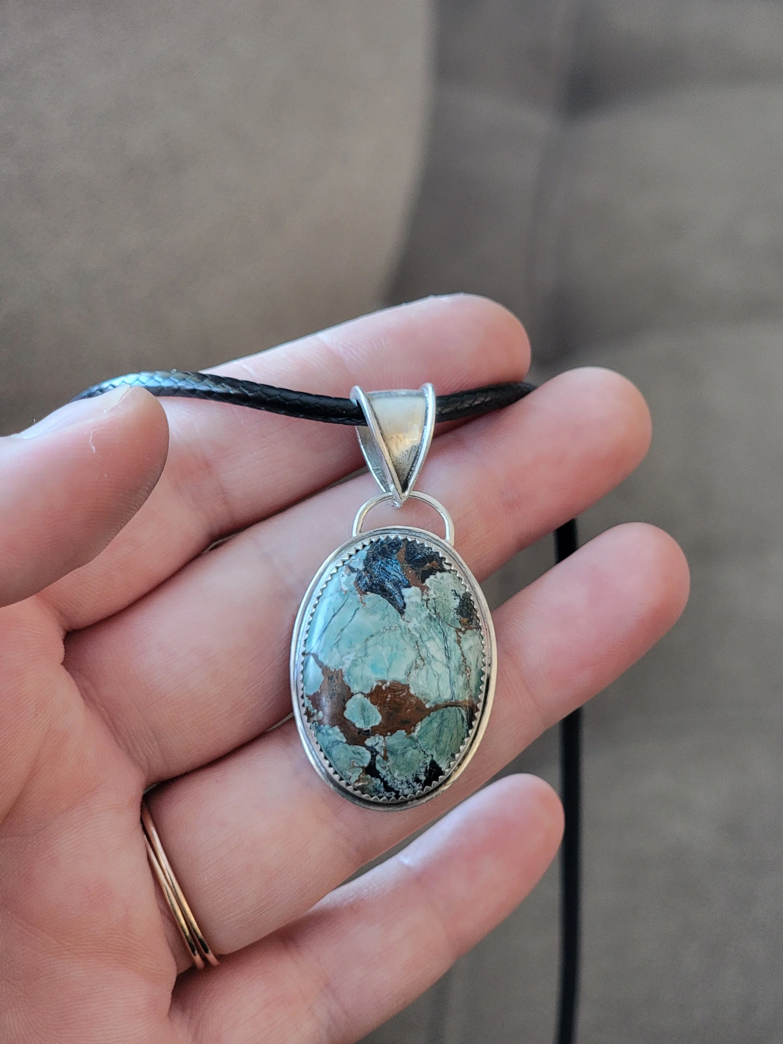 A hand holds a pendant necklace with an oval-shaped stone featuring green, brown, and black patterns, hanging from a black cord.