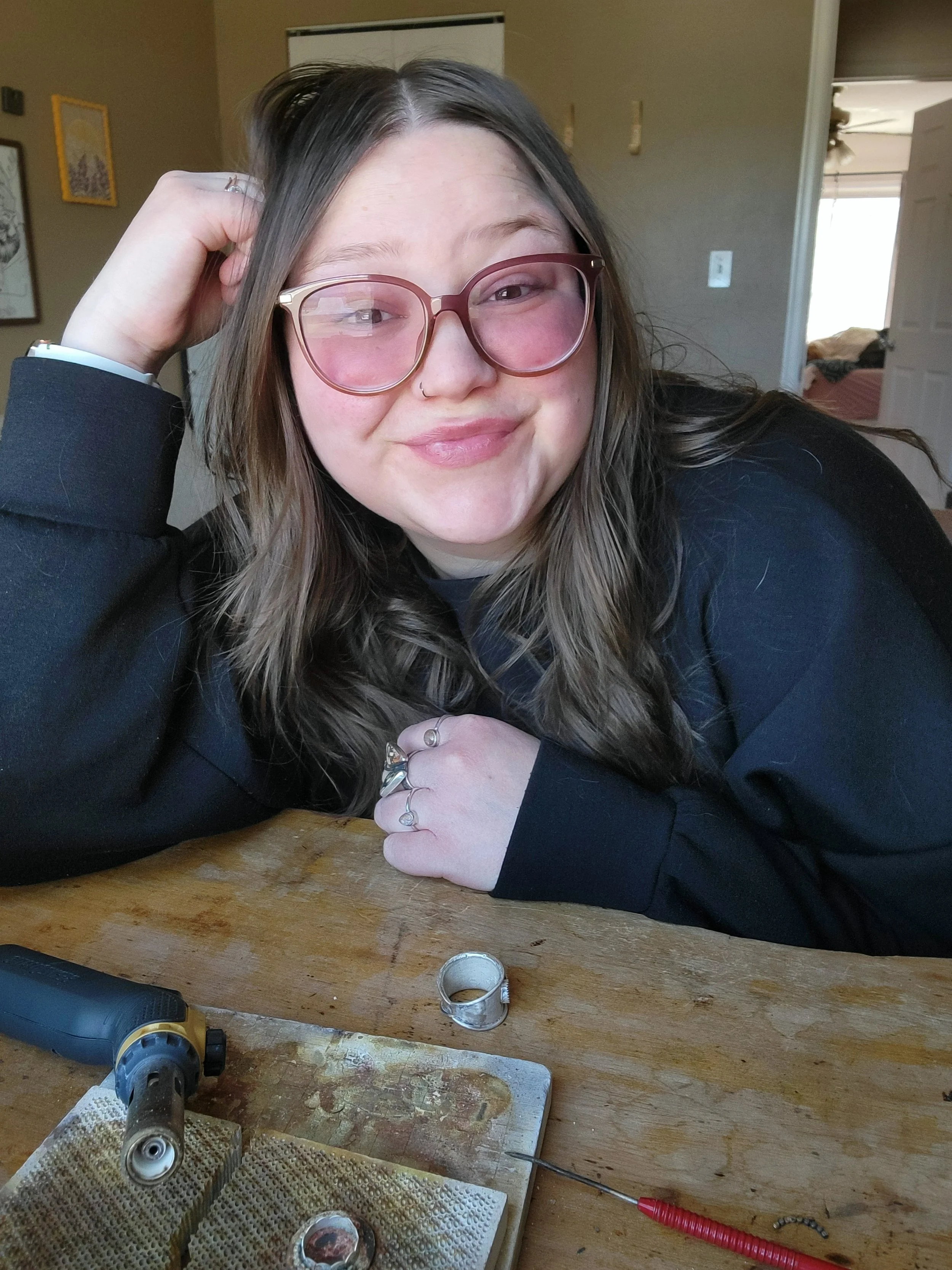 A woman with long brown hair, pink glasses, and a nose ring smiling at the camera, resting her head on her hand. She is sitting at a wooden table with jewelry-making tools and rings in front of her.