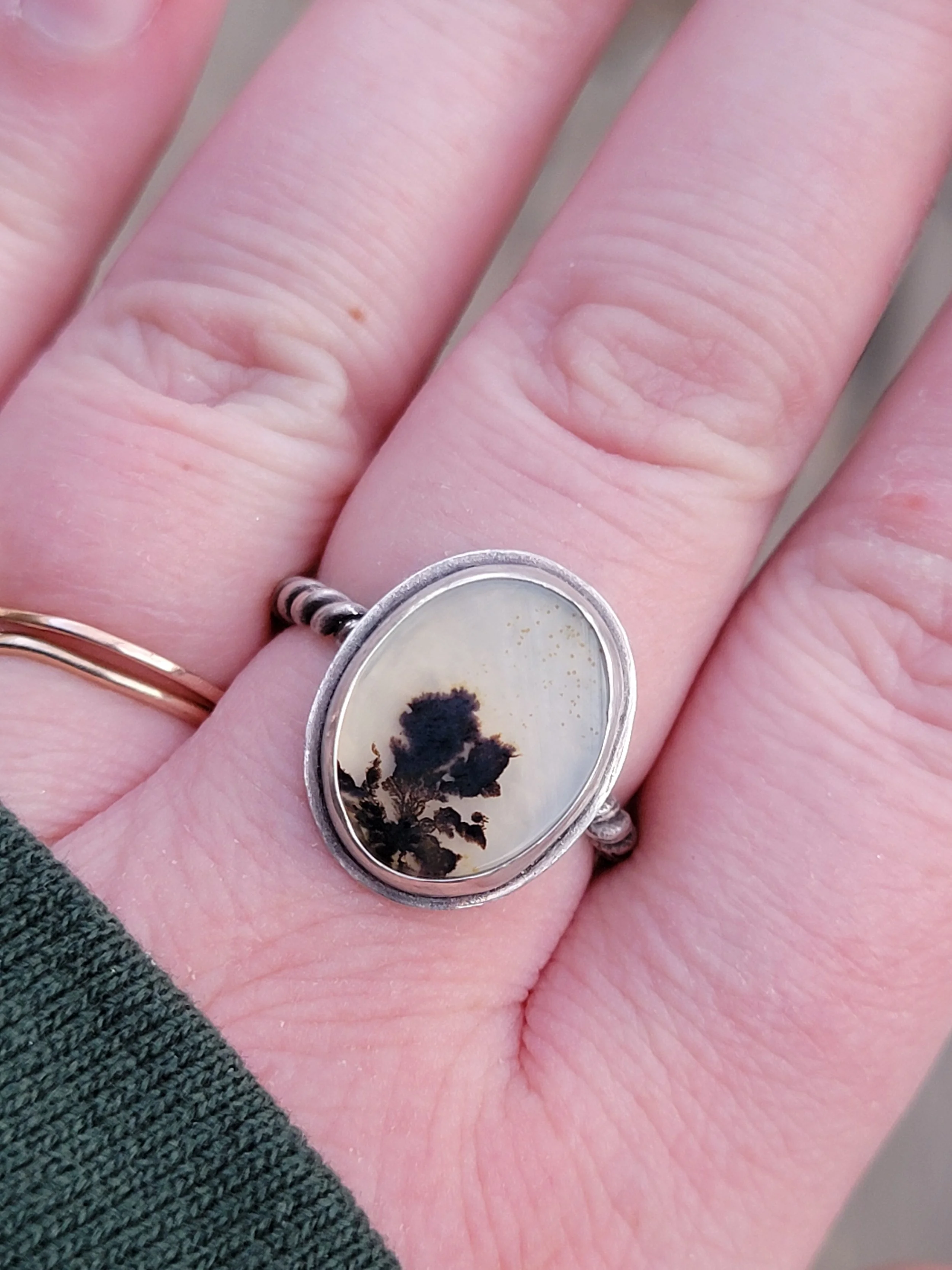A hand with a silver ring featuring an oval-shaped stone containing a black plant silhouette on a light background.