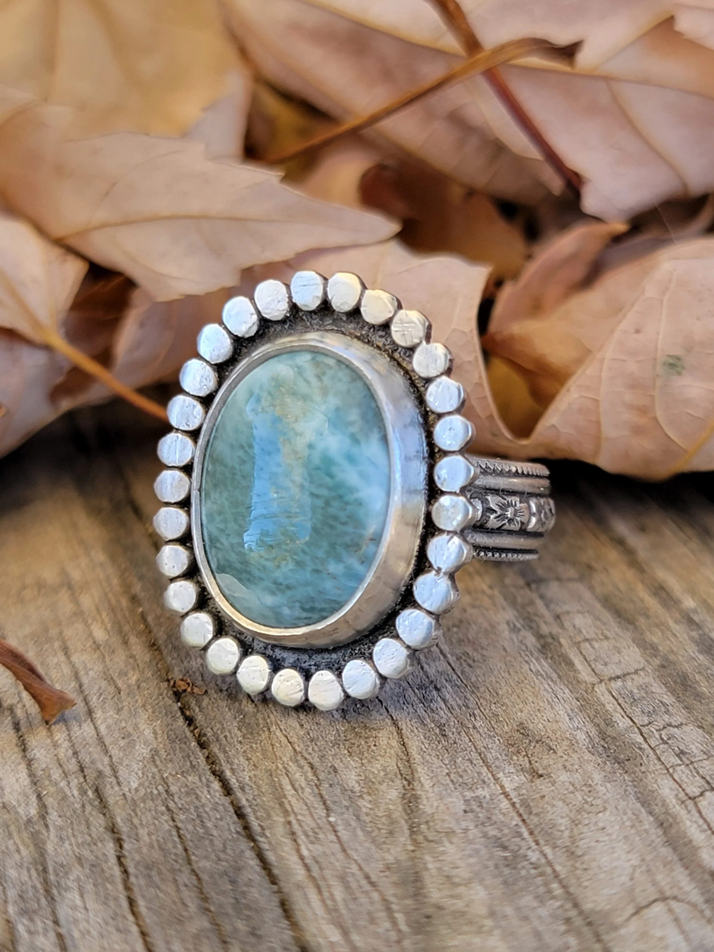 Silver ring with large oval turquoise stone surrounded by small silver beads, placed on a wooden surface with beige dried leaves in the background.