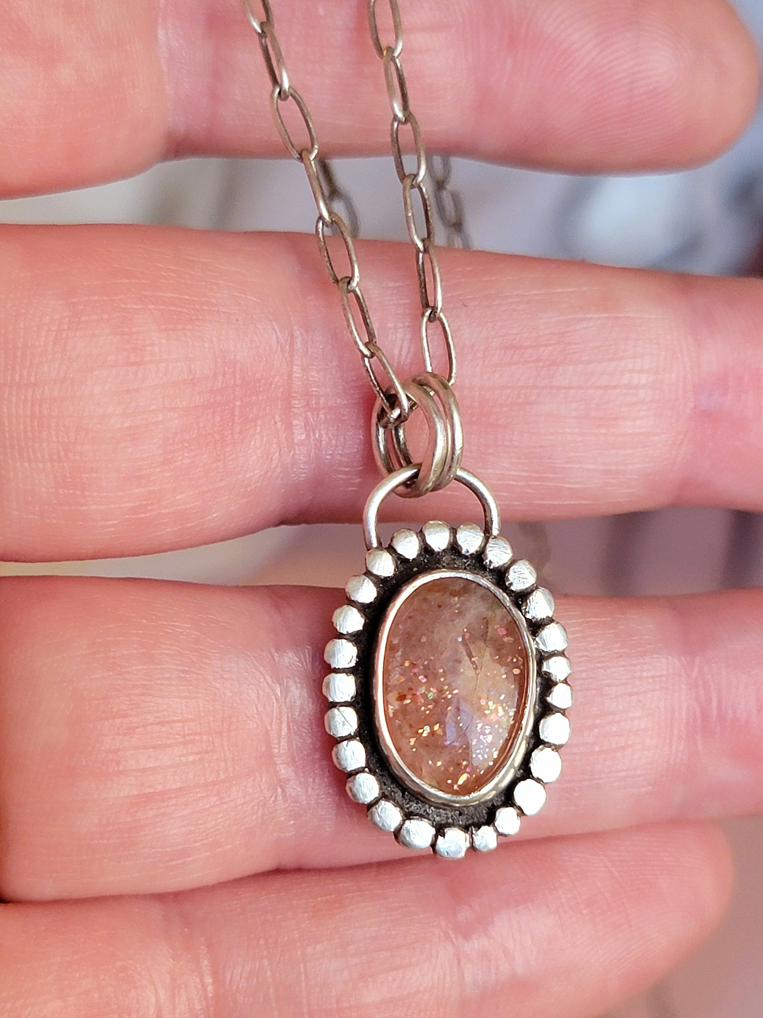 Close-up of a silver necklace with an oval-shaped pendant containing a glittery, iridescent stone, held between fingers.