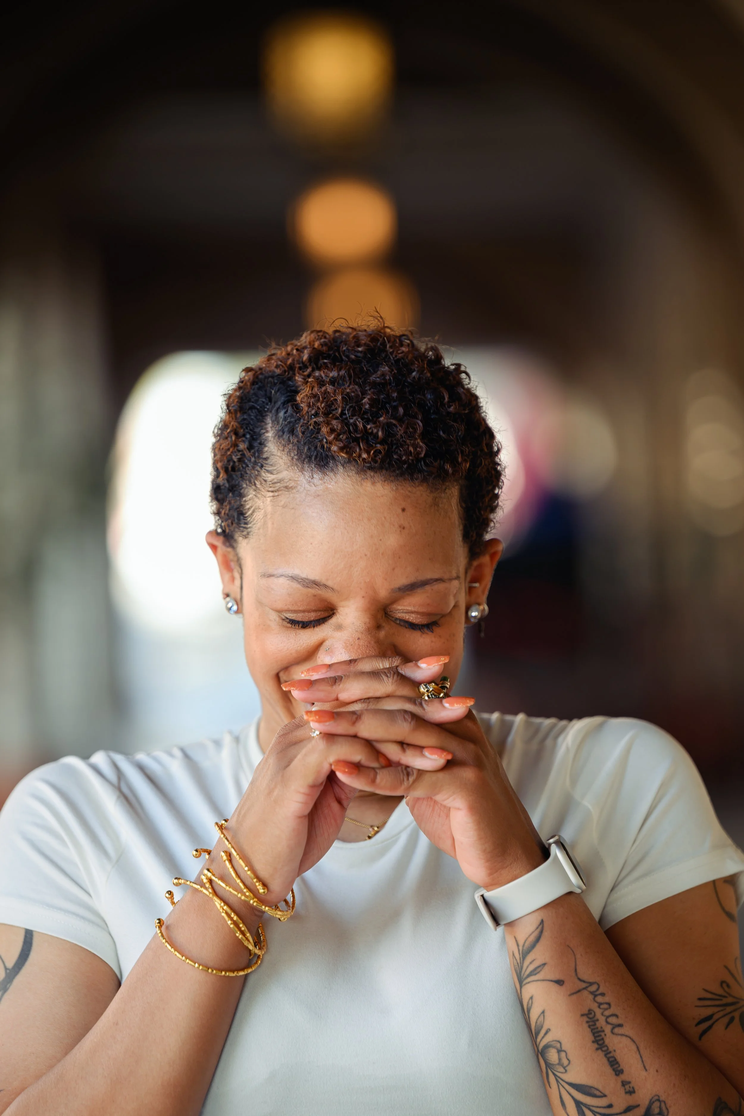 A woman with short, curly hair and multiple tattoos on her arms is smiling with her eyes closed, covering her mouth with her hands while wearing jewelry and a white shirt.