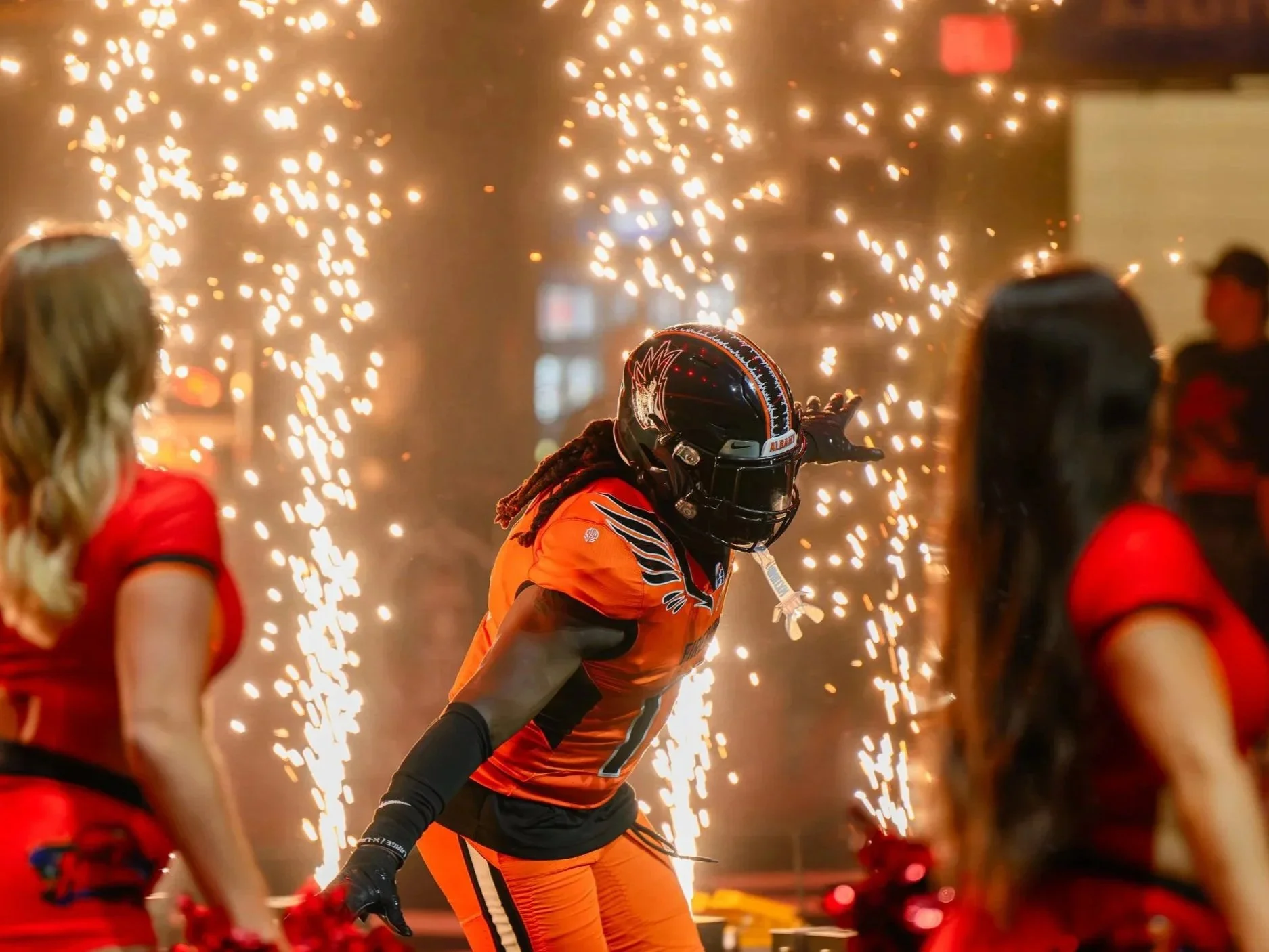 A professional football player in an orange uniform and helmet with a visor is surrounded by sparklers at a celebration event, with two women in red outfits nearby.