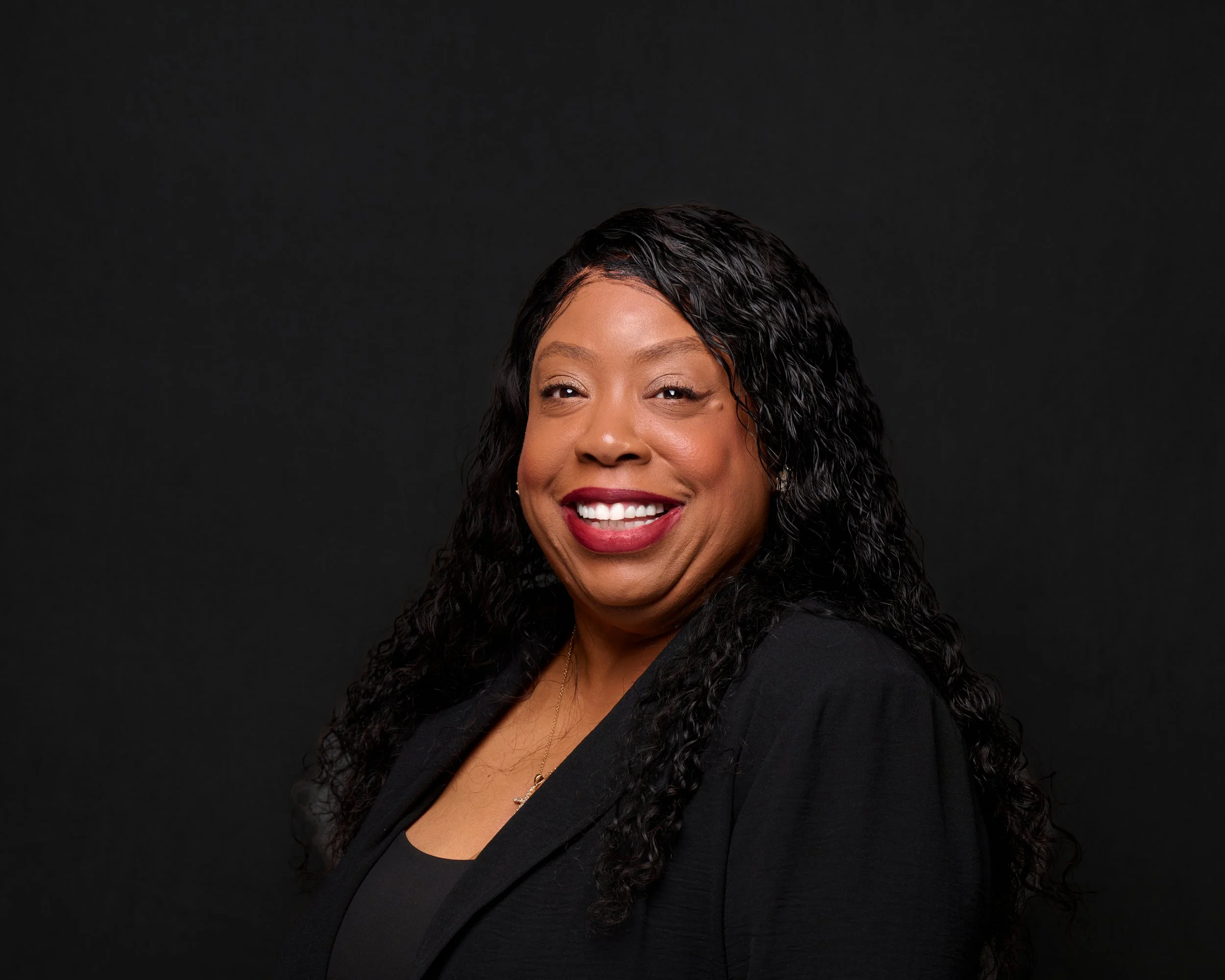 A smiling African American woman with long curly black hair, wearing a black blazer and a black top, against a dark background.