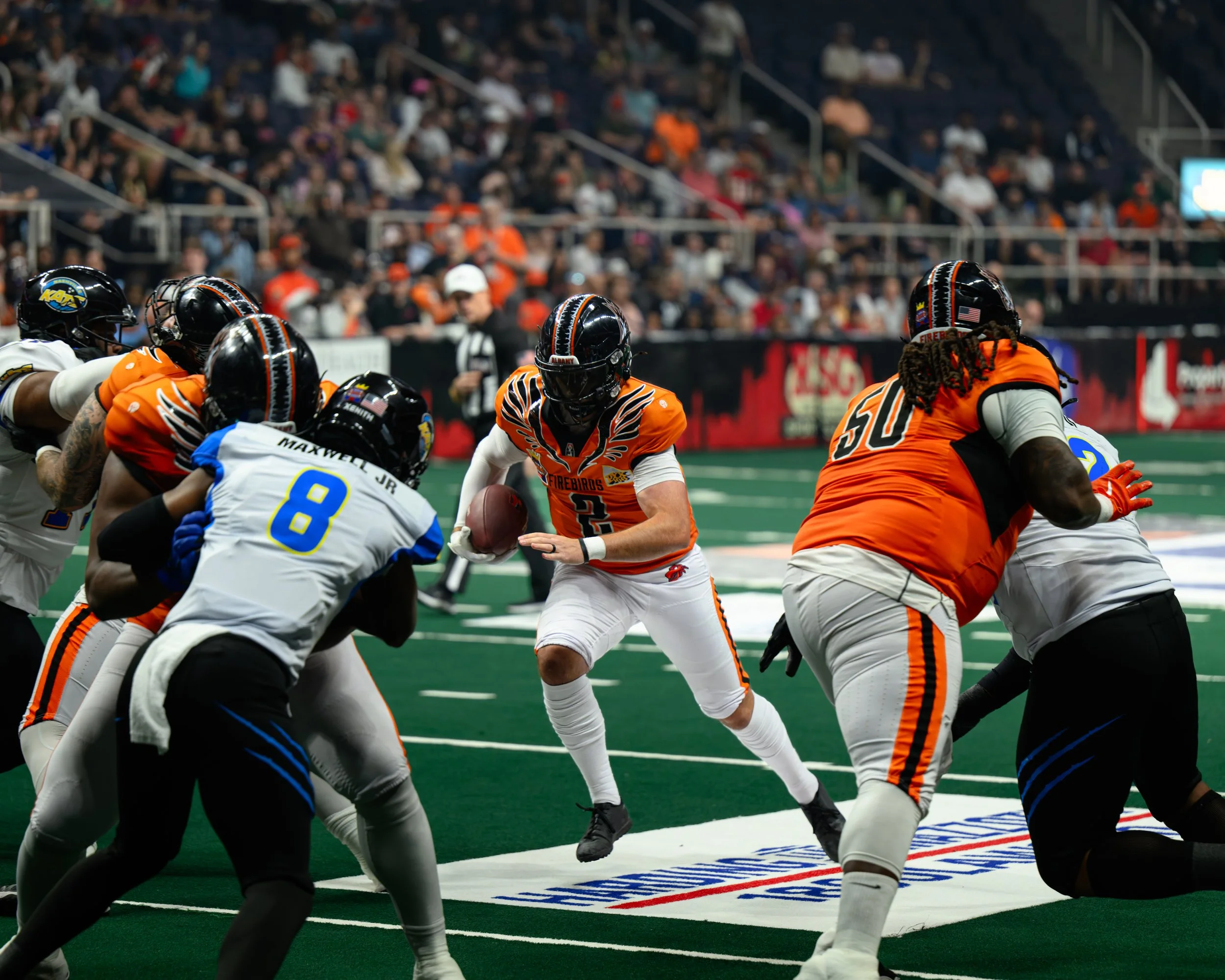 American football players in action on the field during a game, with a player in an orange jersey holding the football and others attempting to block or tackle, in front of a crowd in the stands.