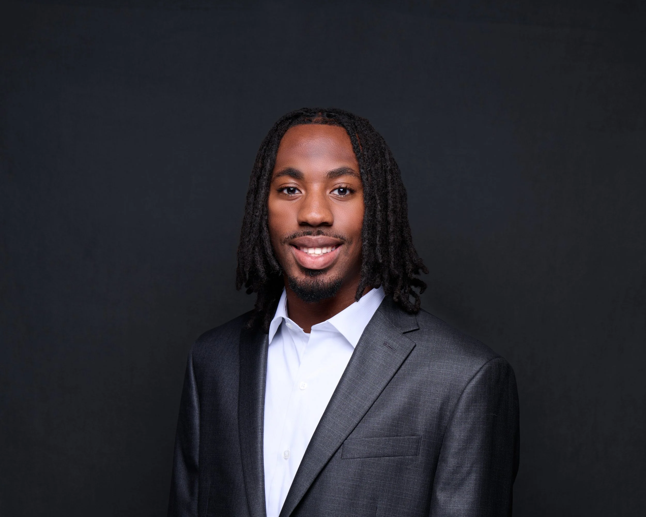 Portrait of a Black man with dreadlocks, wearing a gray suit and white shirt, smiling against a black background.