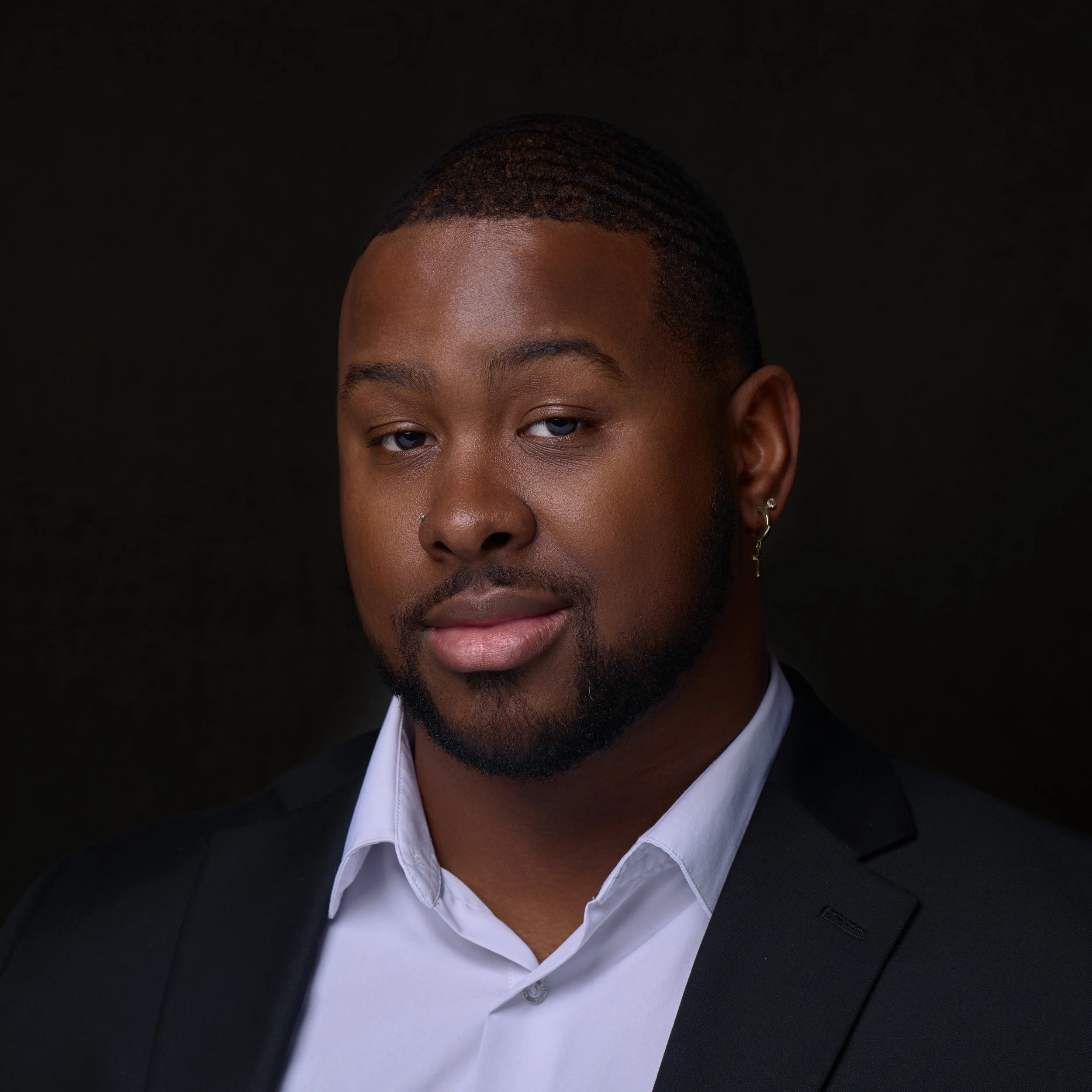 A portrait of a Black man wearing a black suit and white shirt, with short styled hair, a beard, and earrings, against a dark background.