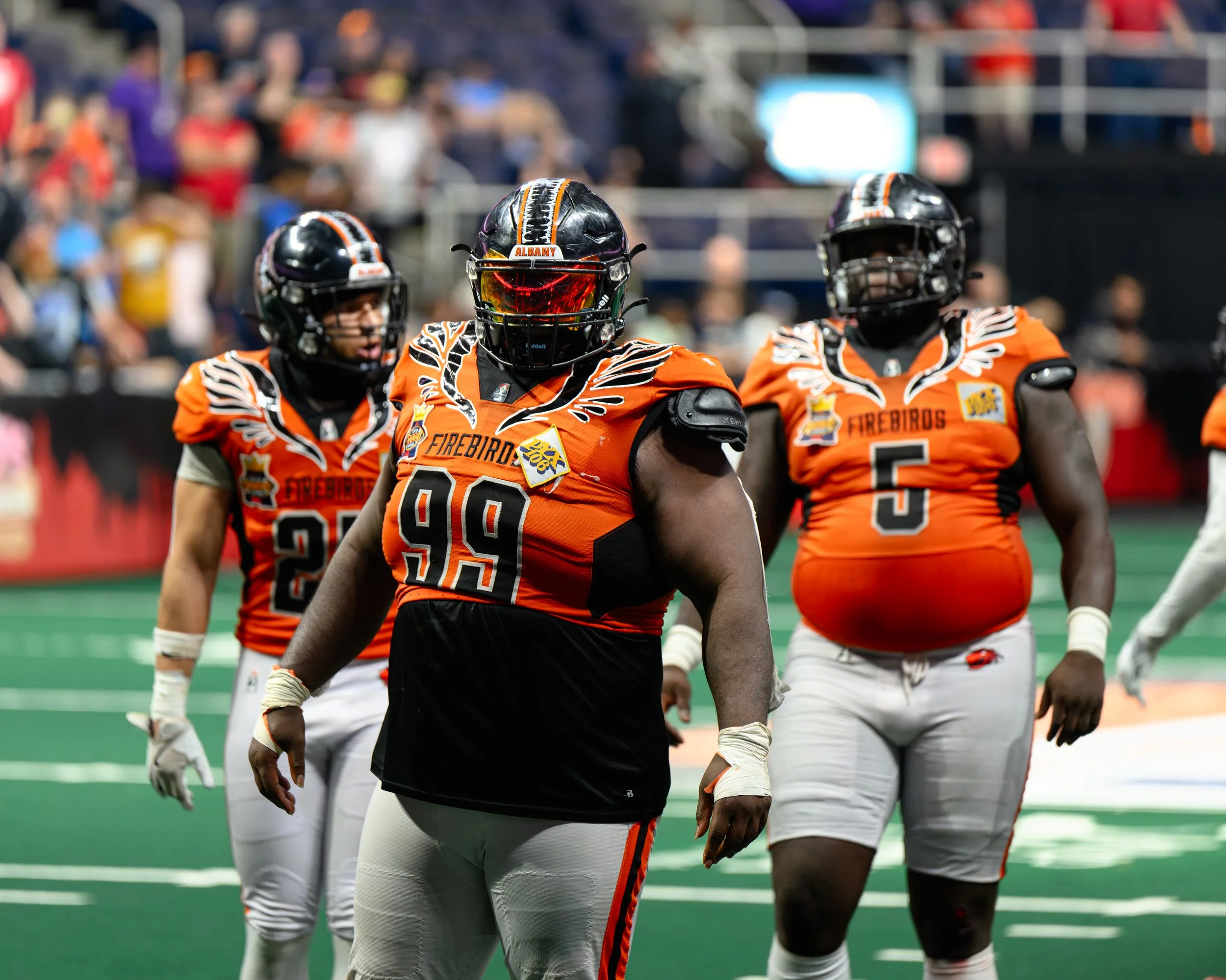 Three football players in orange and black uniforms on the field, with a crowd in the background.