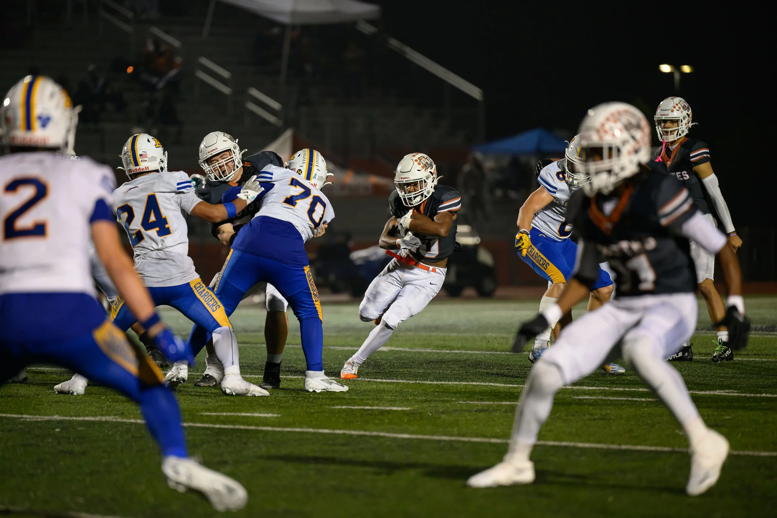 A football game in progress on a field at night with players from two teams, one in black and white uniforms and the other in blue and white uniforms, engaging in a play.