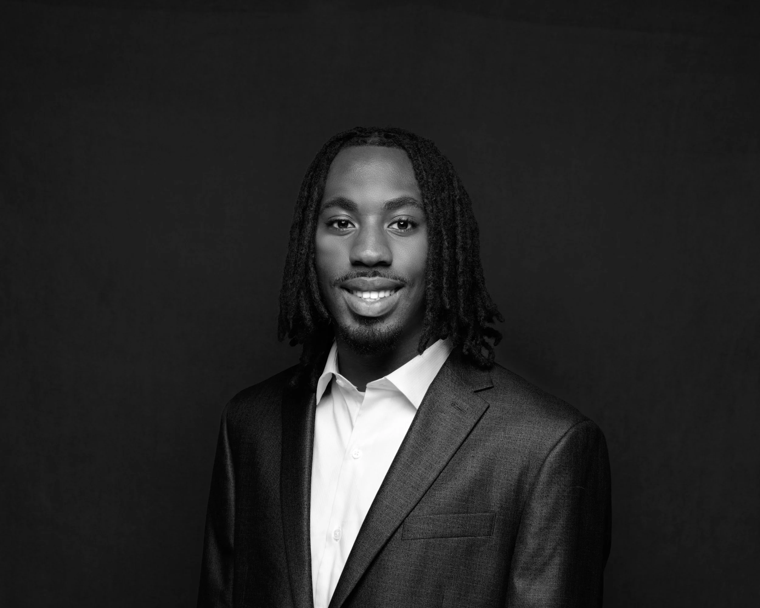 Black and white portrait of a man with dreadlocks, smiling, wearing a suit and white shirt against a dark background.