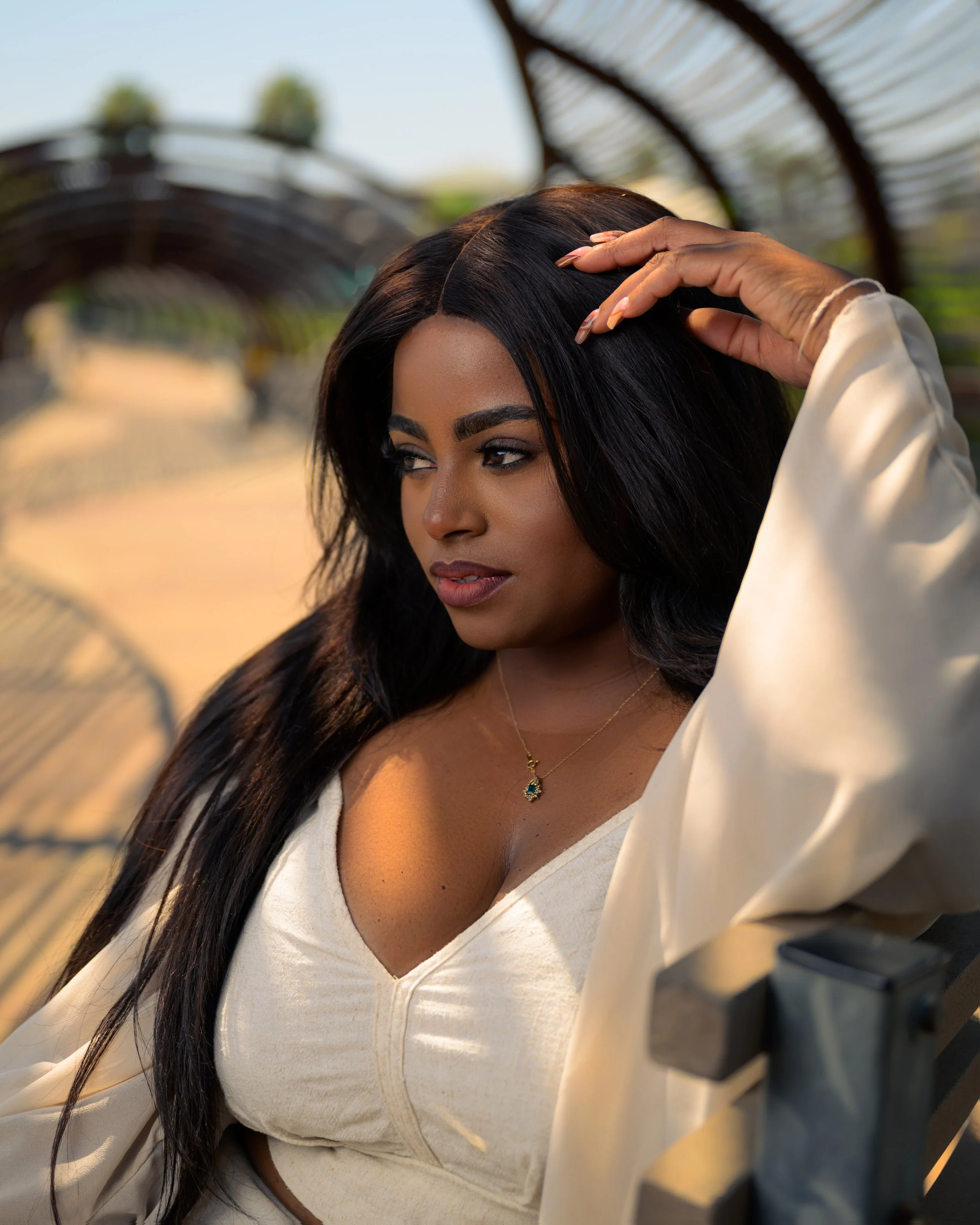 A woman with long black hair and a gold necklace sitting outdoors on a bench, looking pensively to the side, with a blurred park background.