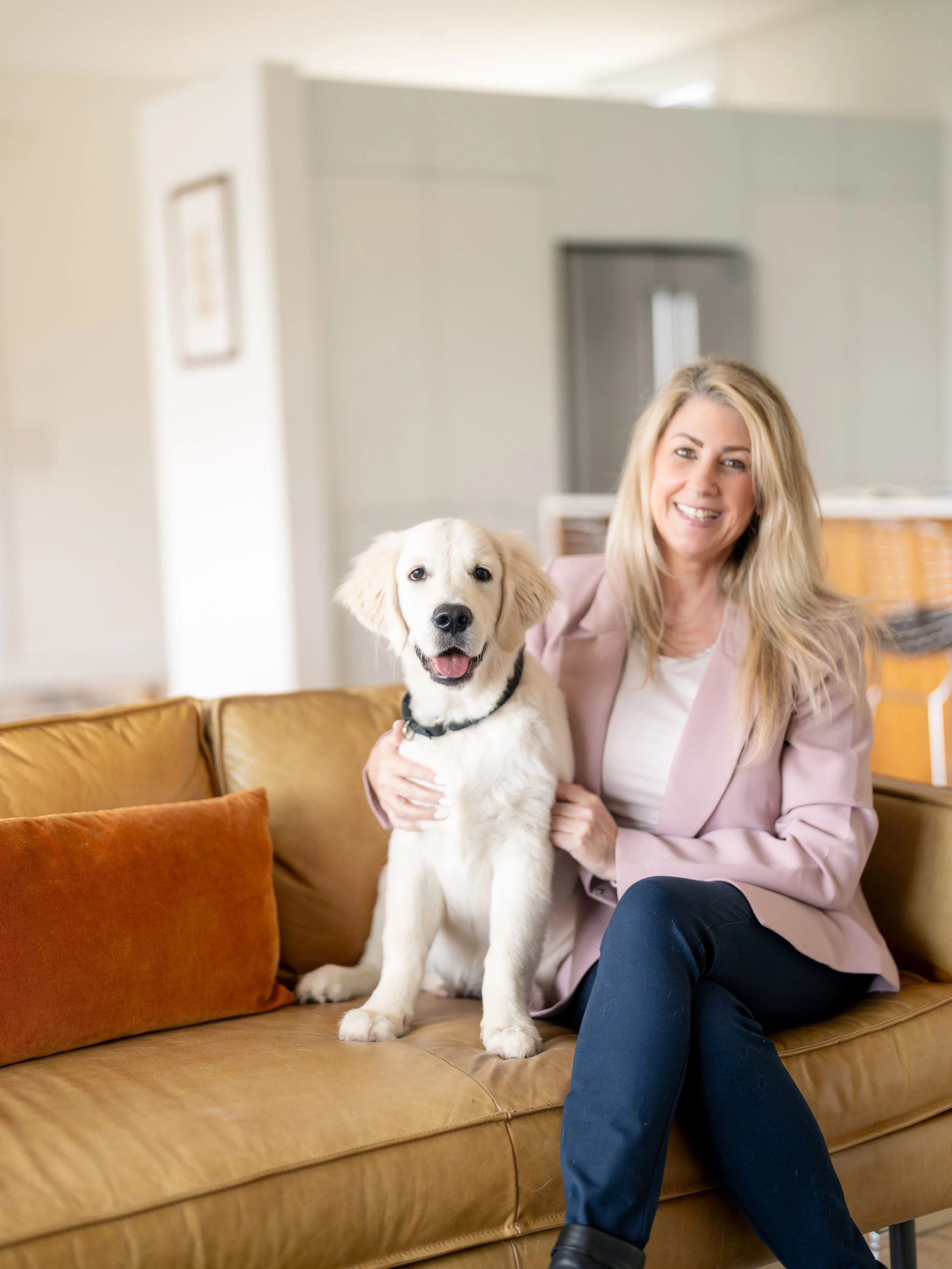 A woman with blonde hair smiling while sitting on a brown leather sofa, holding a white retriever puppy with black eyes and a black collar in a living room.