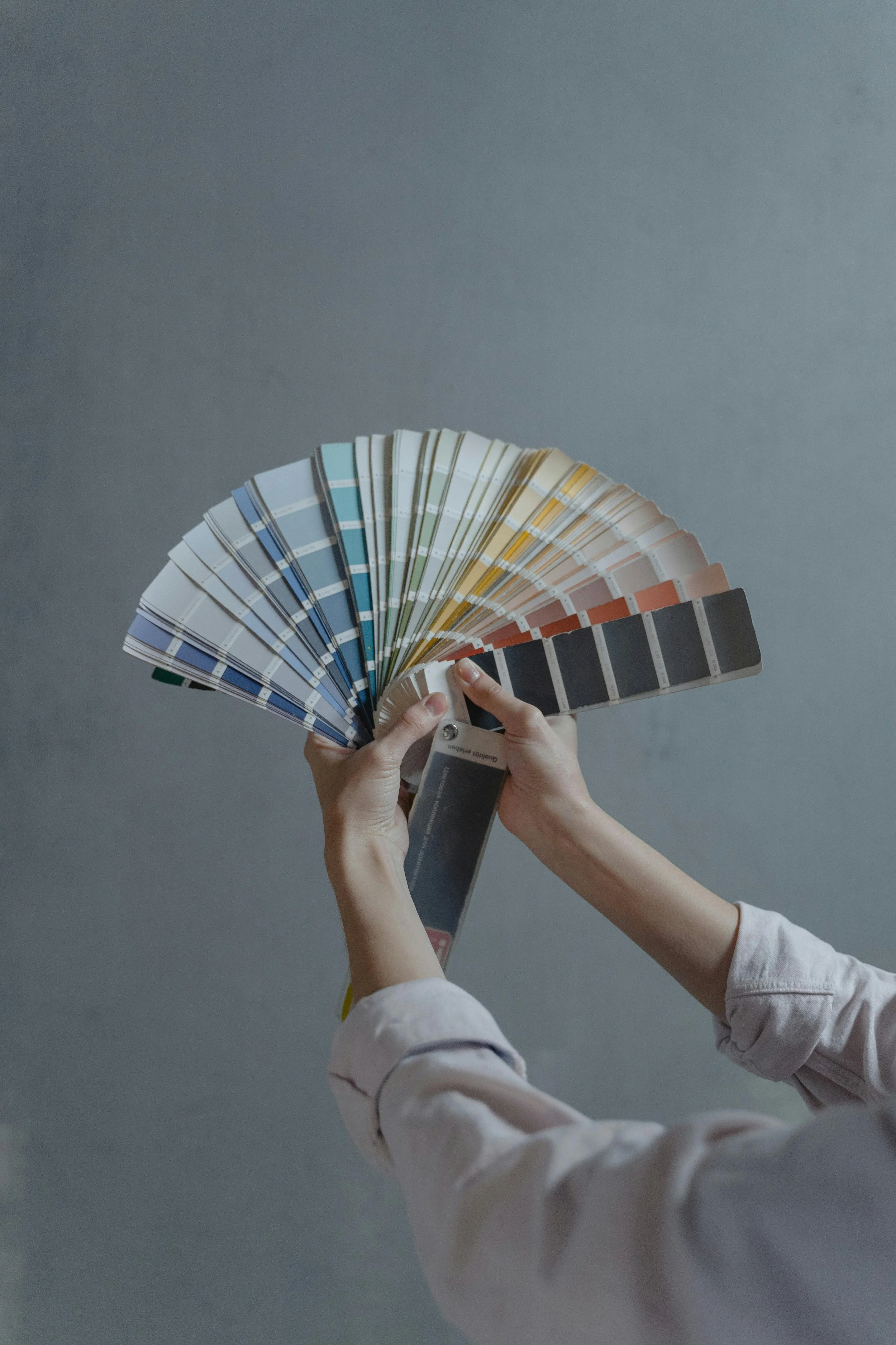A person holding a fan deck of paint color samples with various shades, arranged in a semi-circle, against a plain gray background.