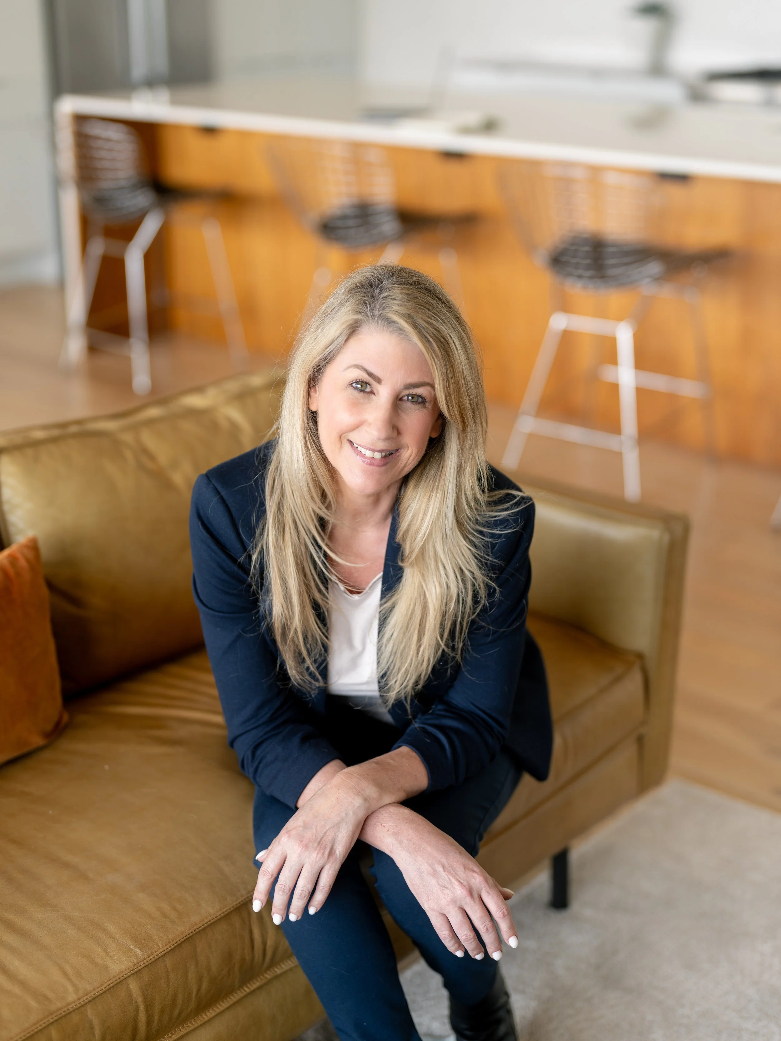 A woman with long blonde hair, wearing a navy blazer and white top, sitting on a tan leather sofa in a modern room with a kitchen in the background.