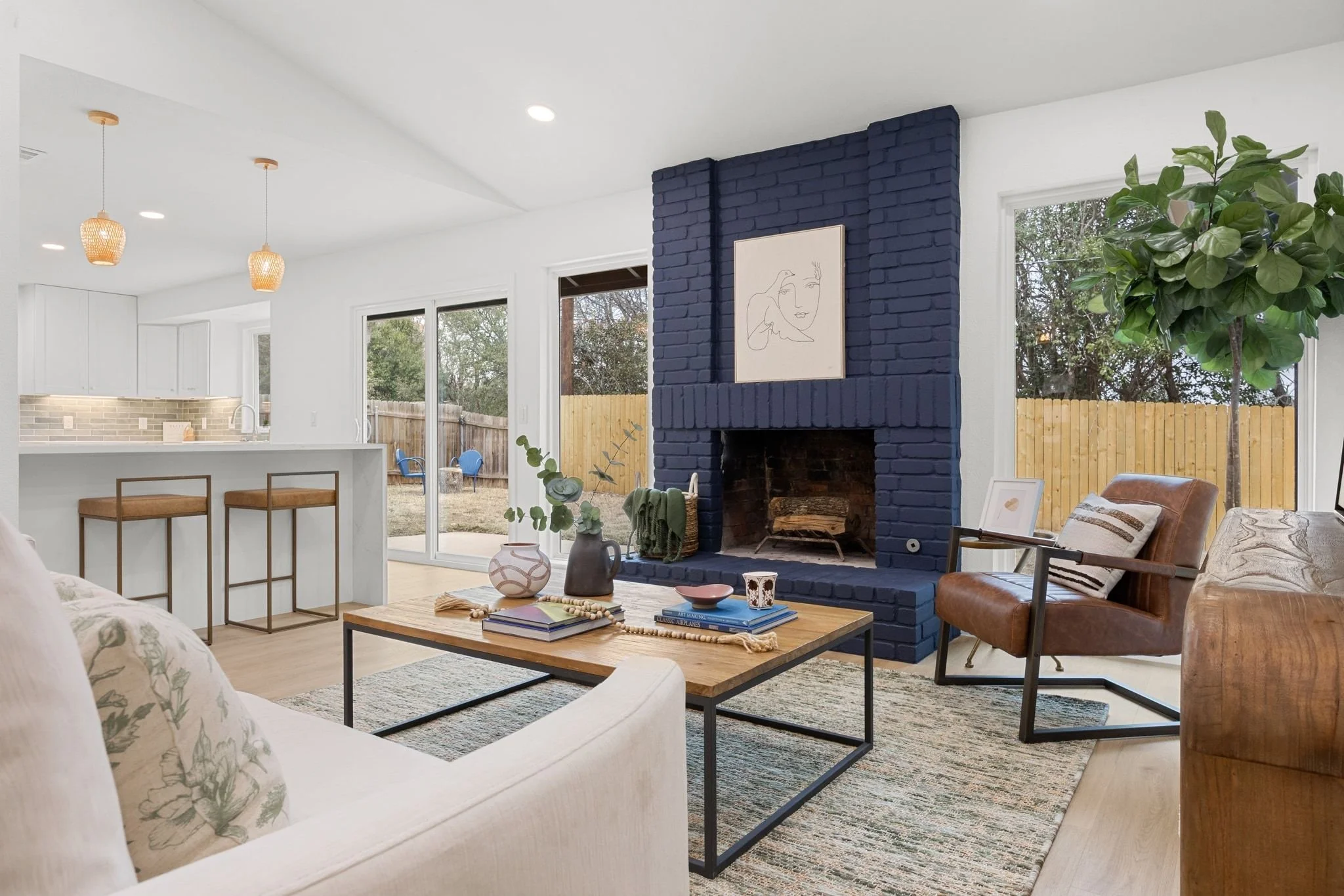 Living room with blue brick fireplace, white walls, sliding glass doors, a wooden coffee table with books and decor, leather and floral sofas, indoor plants, and decorative lighting fixtures.