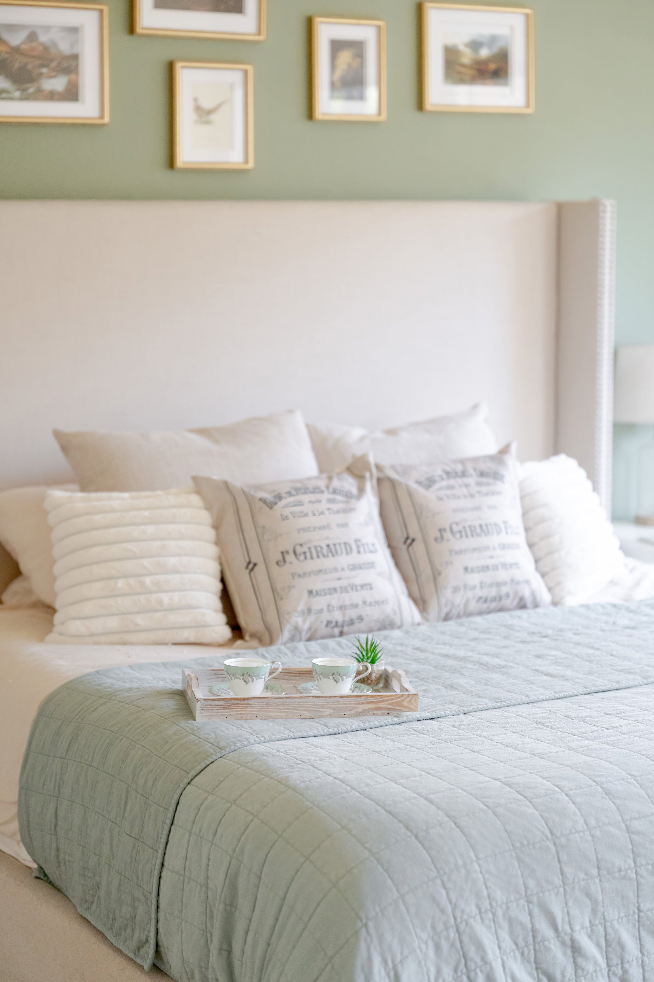 A neatly made bed with a light green quilt, a variety of cream-colored pillows, and a tray with two teacups. The headboard is tall, beige, and upholstered. The background features a light green wall decorated with several framed pictures.