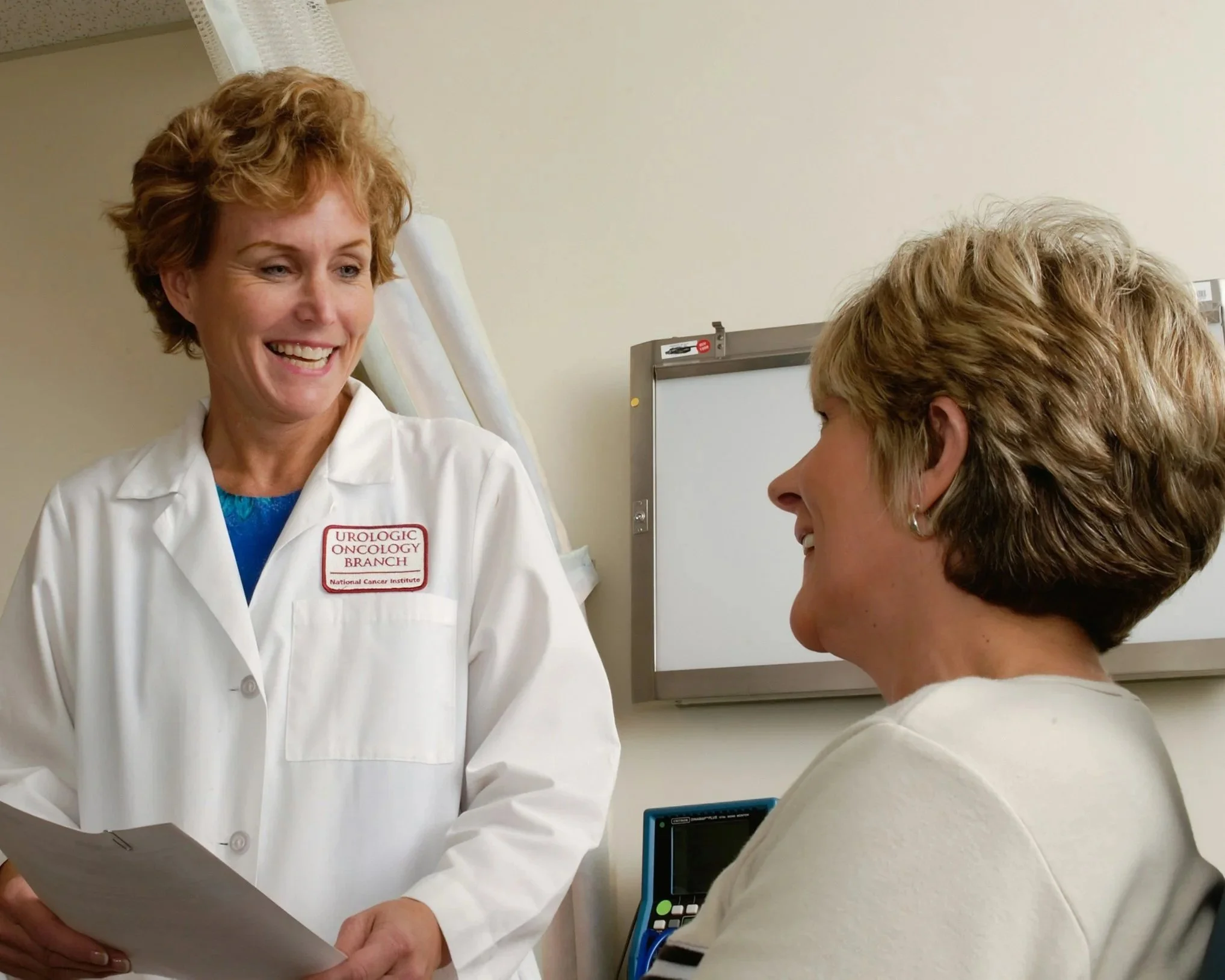 A female doctor and female patient having a conversation in a medical office, with the doctor smiling and holding a clipboard.