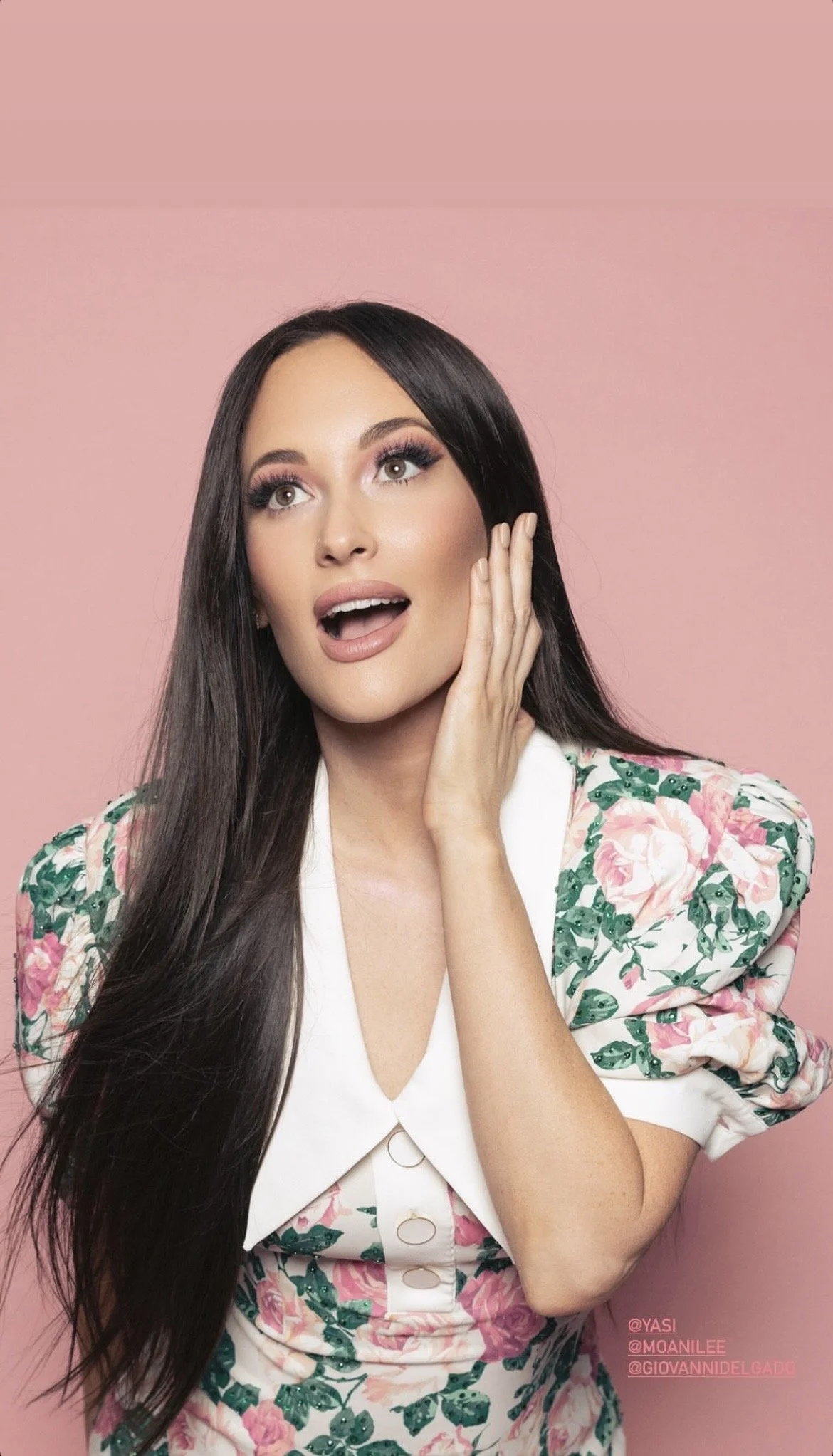 A woman with long dark hair, makeup, and a surprised expression, touching her face against a pink background. She is wearing a white blouse with a floral patterned jacket.