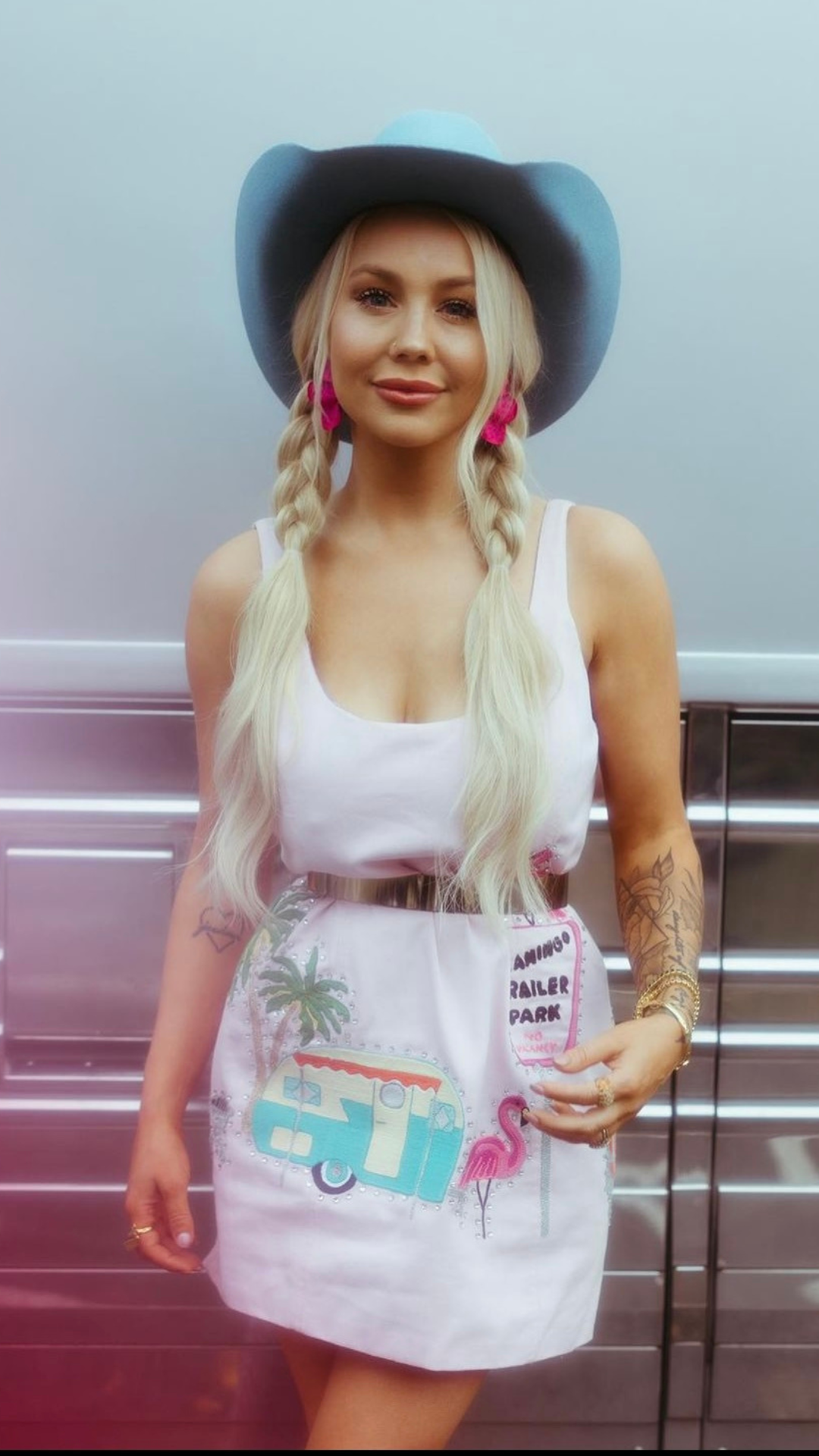 A woman with long blonde hair in braids wearing a large blue hat, white tank top, and a white apron with colorful designs, standing in front of an elevator.