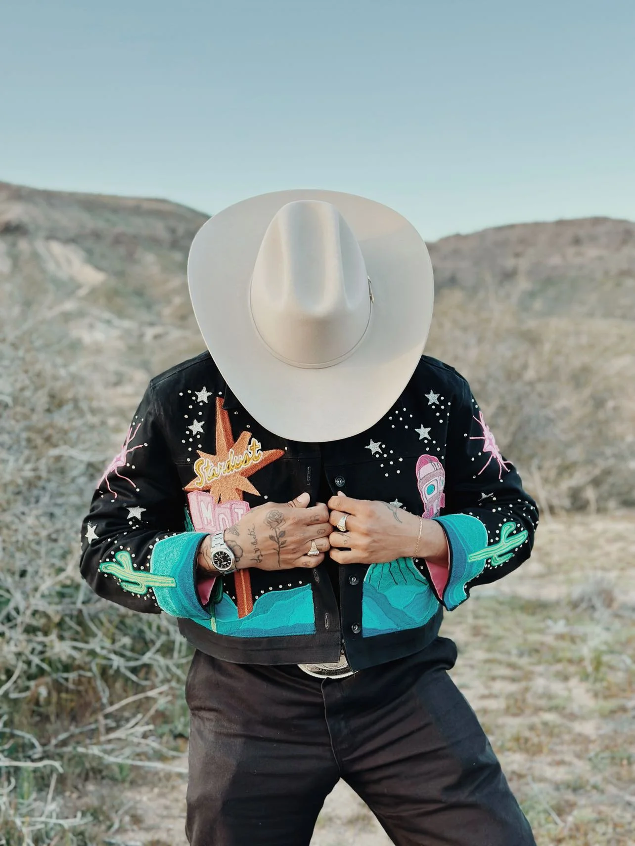 A person standing outdoors in a desert-like landscape, wearing a white cowboy hat and a black embroidered jacket, holding the jacket closed, with tattoos and jewelry visible, and mountains in the background.