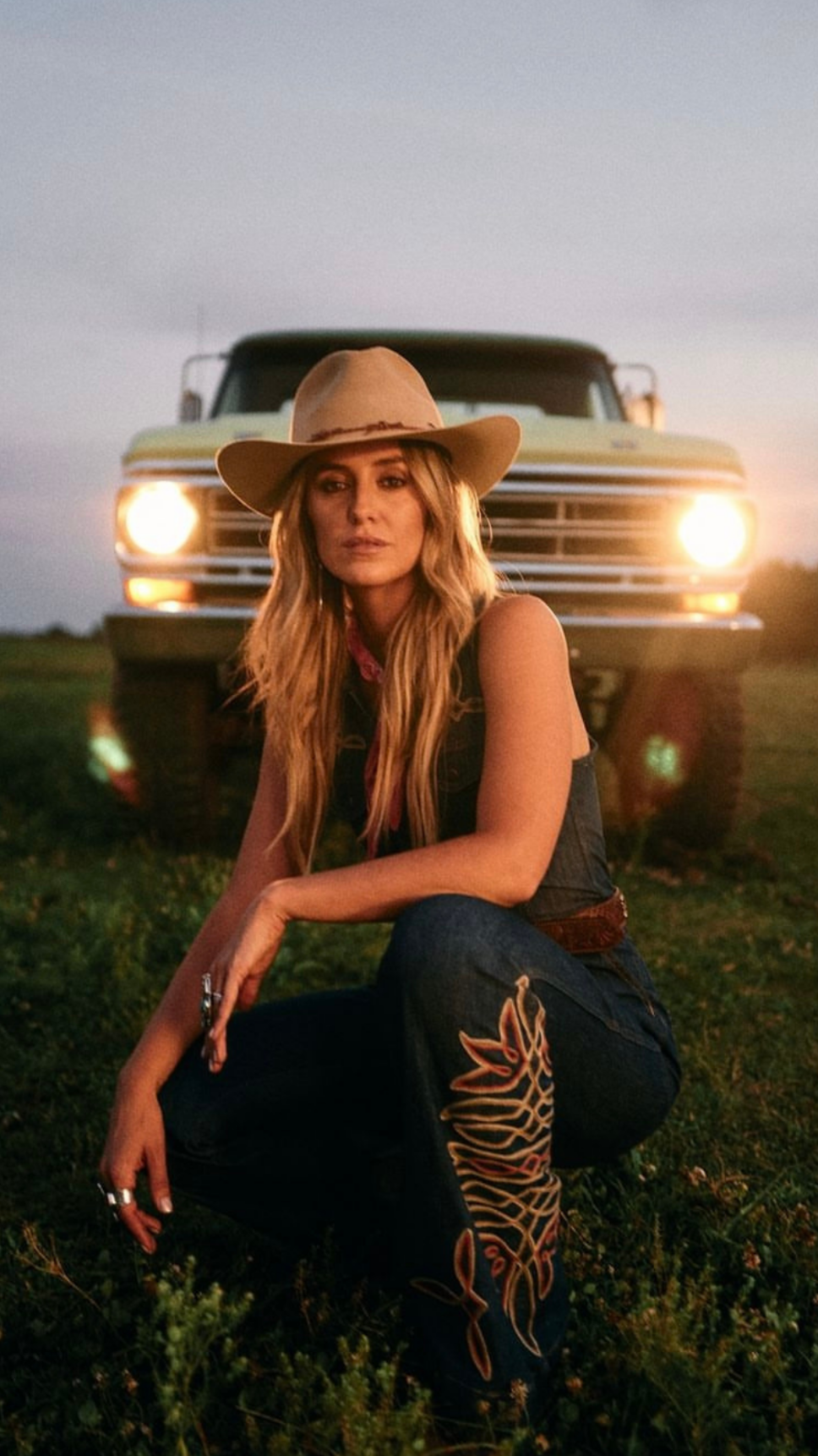 A woman with long blonde hair wearing a cowboy hat, denim vest, and embroidered jeans, squatting on grass during sunset with an old yellow truck with headlights on behind her.
