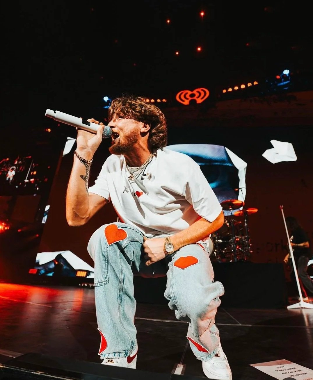 A male singer performing on stage, kneeling with one knee up, holding a microphone close to his mouth, wearing a white t-shirt with a heart graphic, distressed jeans with red patches, and jewelry, with a band and large screen in the background.