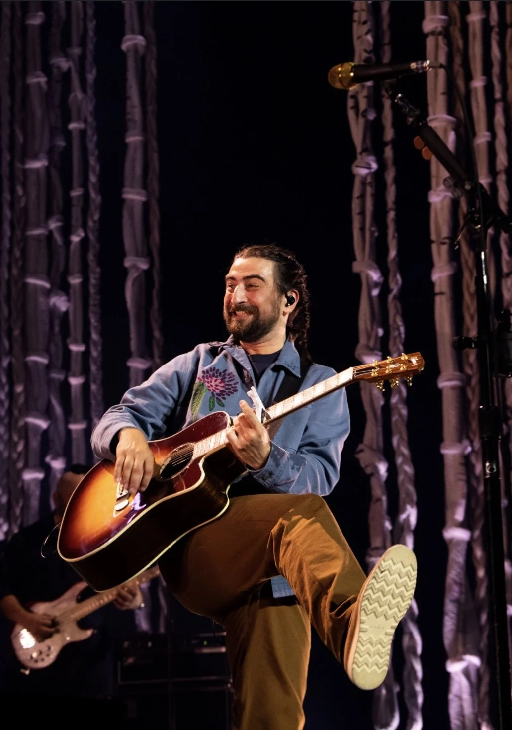 A musician with a beard and long hair tied back is performing on stage with an acoustic guitar, smiling and wearing a blue shirt with floral embroidery, brown pants, and in-ear monitors. In the background, another musician is playing a bass guitar, a