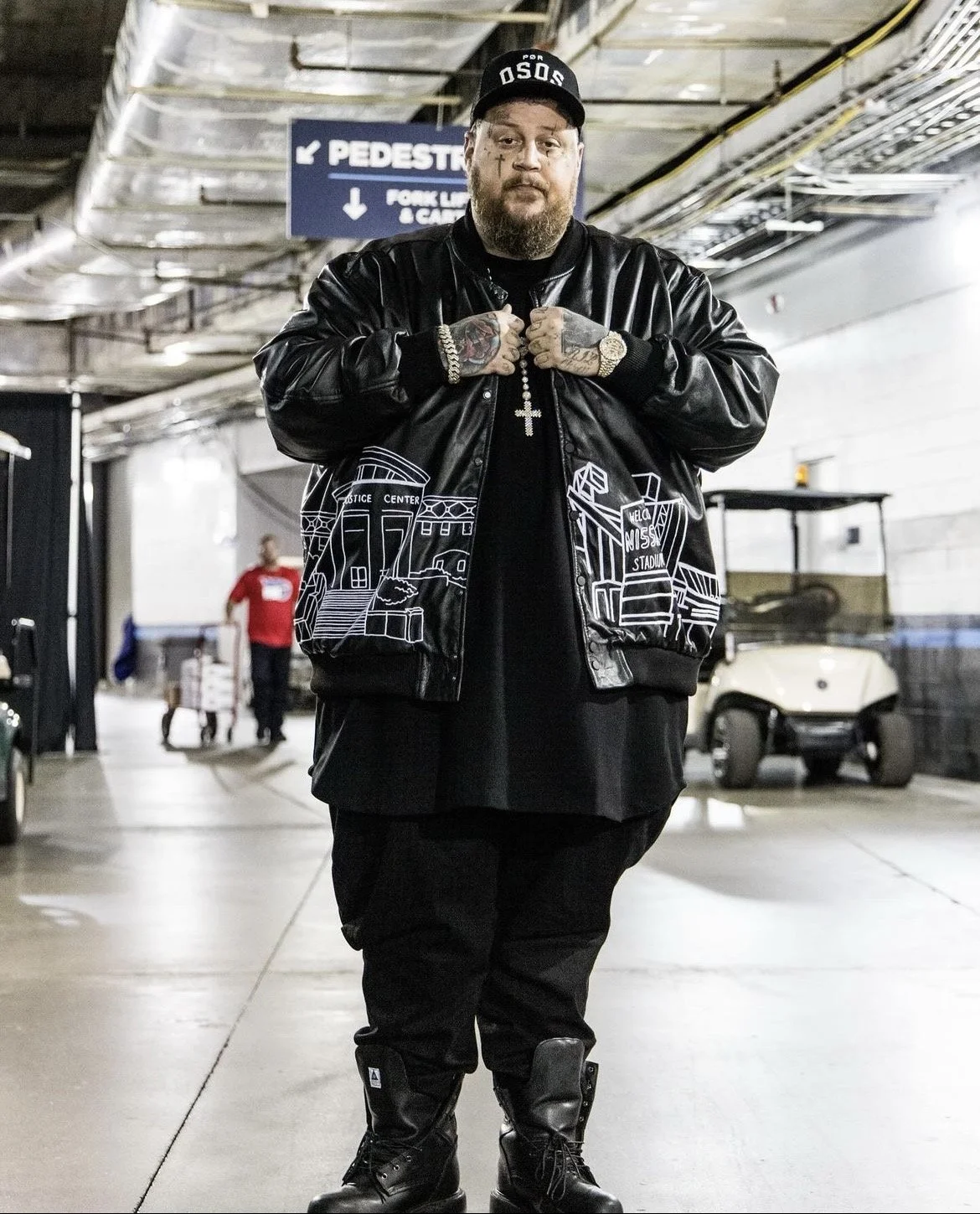 A man in black clothing with tattoos, jewelry, and a black cap stands in a parking garage, with a golf cart and a person in red in the background.
