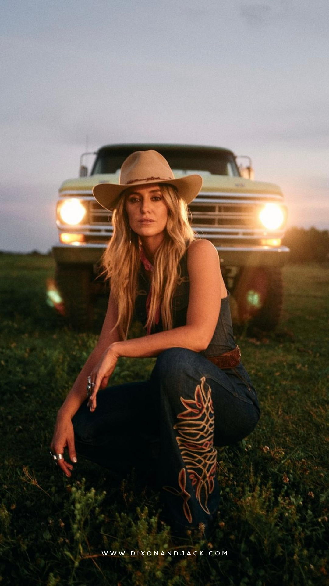 A woman with long blonde hair wearing a cowboy hat, black sleeveless shirt, and embroidered jeans, sitting on the grass in front of a yellow truck at sunset.